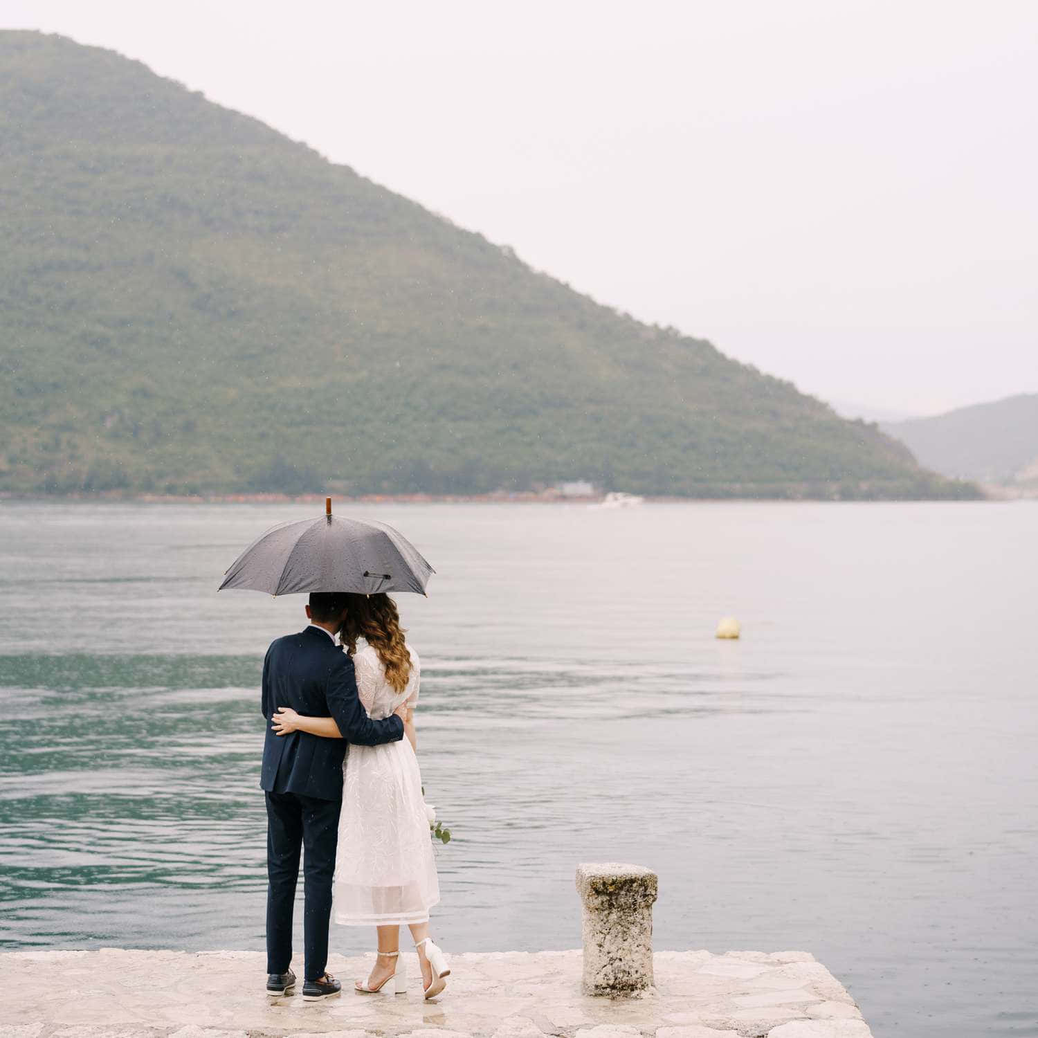 Dock 桌布 壁紙 — 一對情侶在石碼頭上撐著灰色雨傘擁抱，俯瞰平靜的大海，背景是霧氣瀰漫的綠色山丘。
