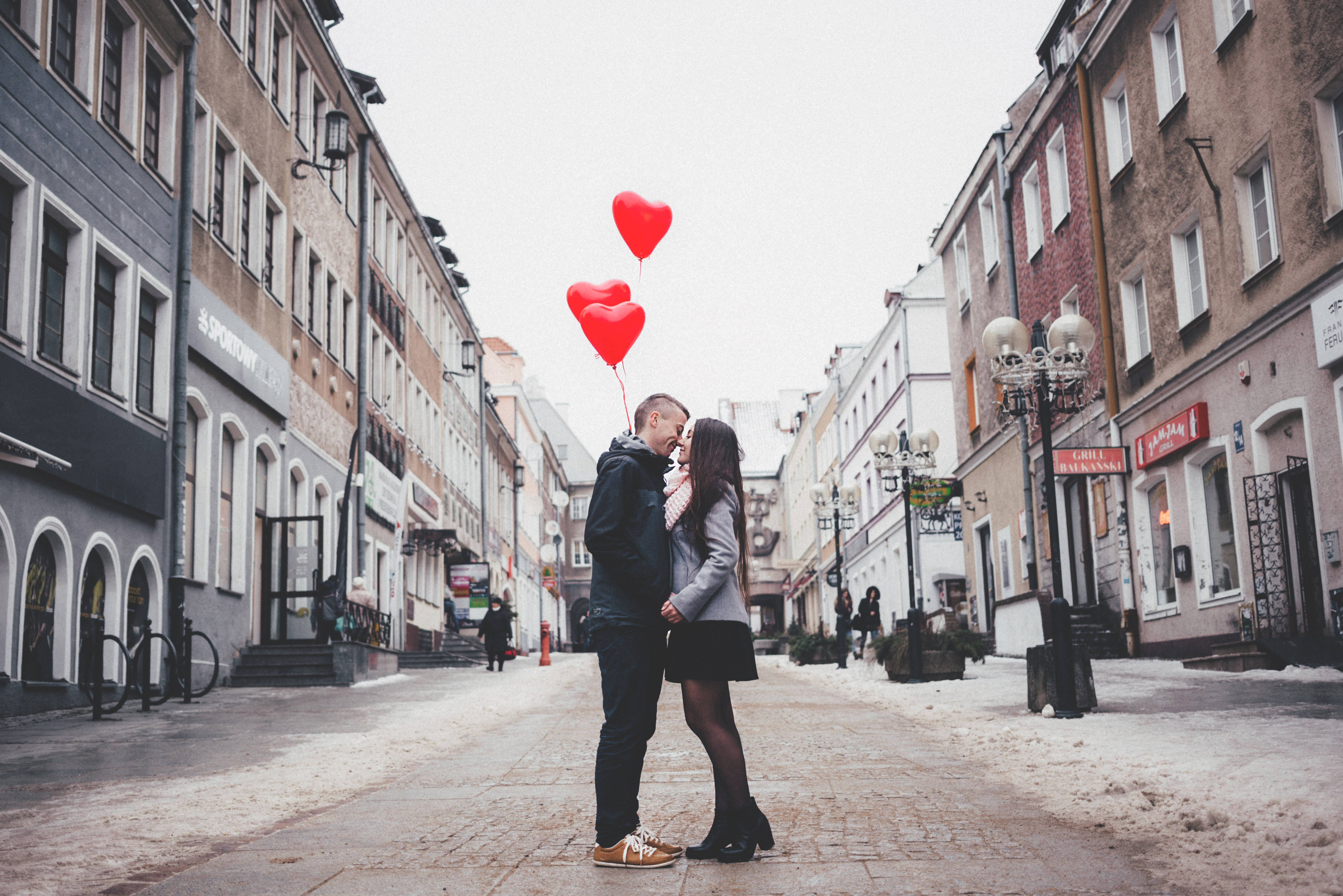 couple kiss 桌布 壁紙 — 一對情侶在積雪的城市街道上接吻，手持紅色心形氣球，背景是灰色建築物。