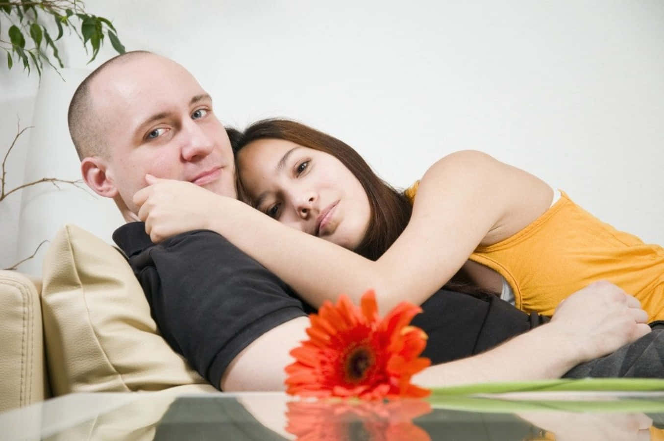 Sofa 桌布 壁紙 — 一對情侶在米色沙發上親密擁抱，前景有一朵鮮豔的橙色非洲菊雛菊。