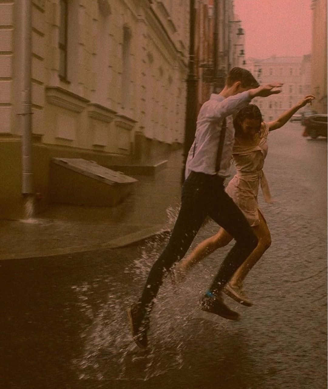 Aesthetic Couple 桌布 壁紙 — 一對情侶在粉紅色的天空下，歡快地跳過濕滑的都市小巷，濺起水花。
