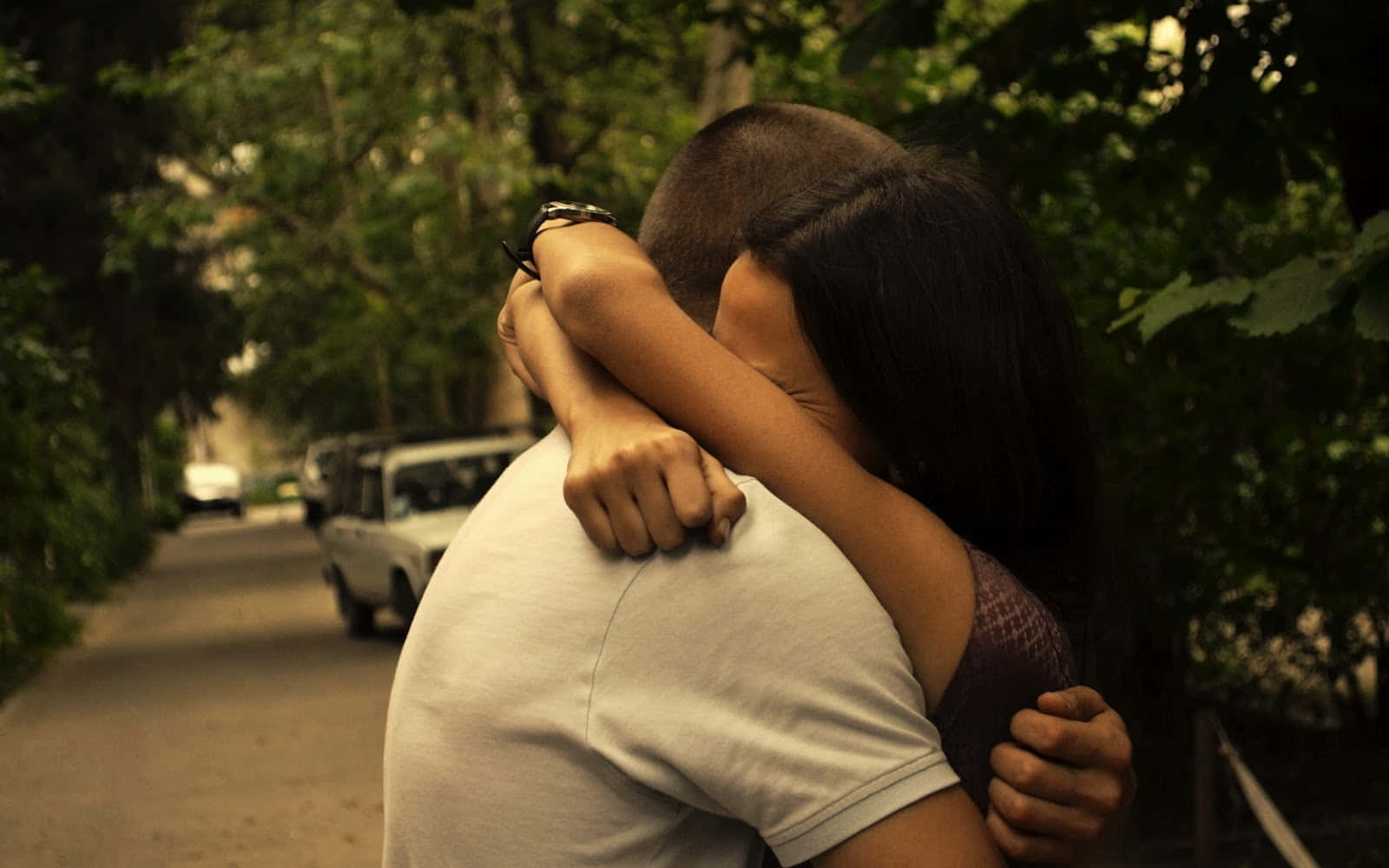 Sad Couple 桌布 壁紙 — 一對情侶在綠樹成蔭的街道上溫暖地擁抱，附近停有復古汽車。