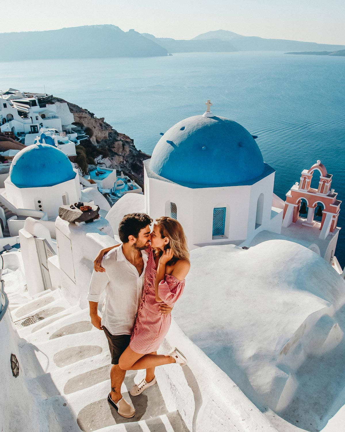 romantic kiss 桌布 壁紙 — 一對情侶在聖托里尼的白色階梯上接吻，旁邊是藍頂教堂，俯瞰愛琴海。