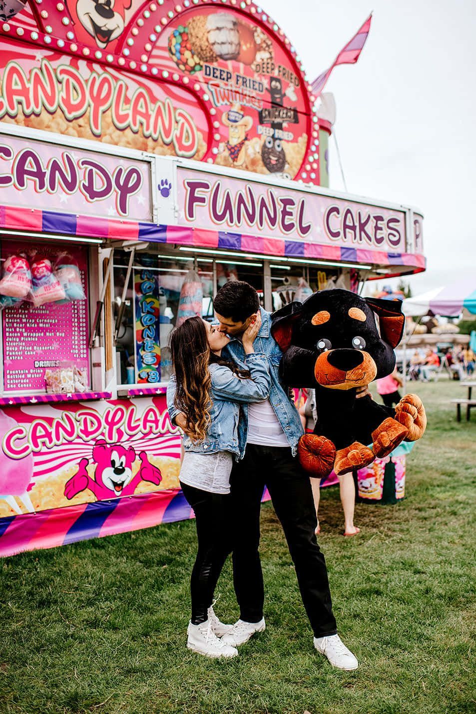 Carnival 桌布 壁紙 — 一對情侶在色彩繽紛的糖果樂園美食車前接吻，車上有一個巨大的毛絨狗吉祥物拿著漏斗蛋糕。