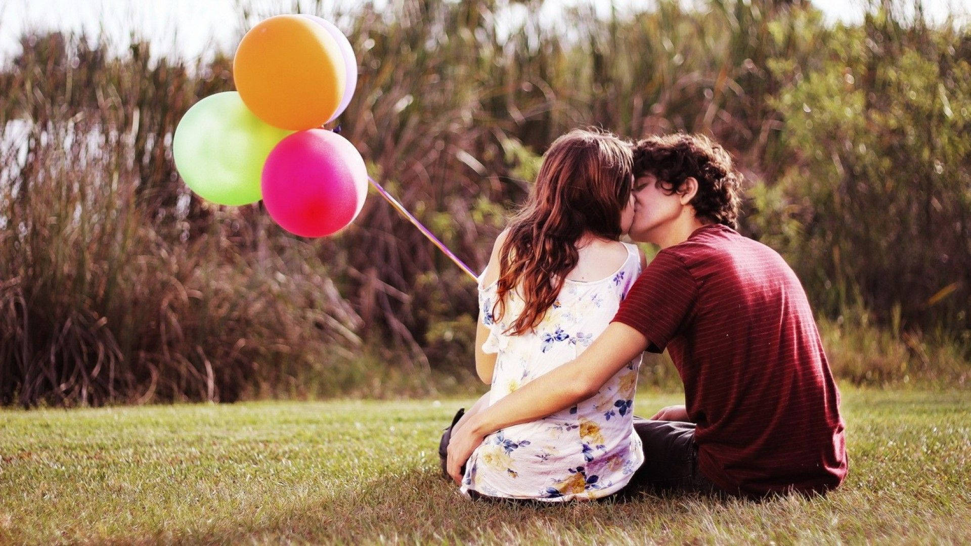 couples kissing 桌布 壁紙 — 一對情侶在草地上接吻，手持彩色氣球，背景是模糊的自然景色。