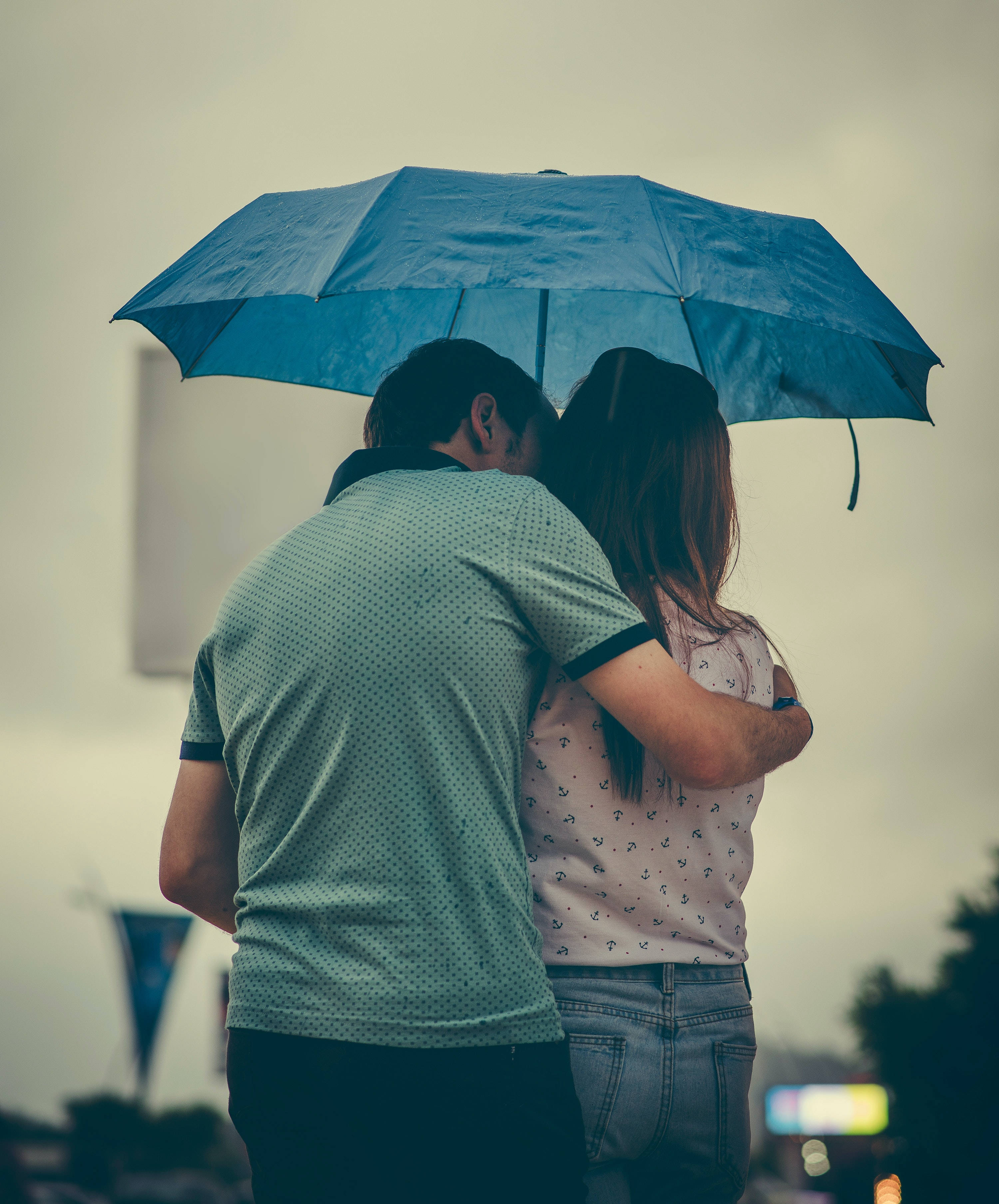 Love Cute Couple 桌布 壁紙 — 一對情侶在藍色雨傘下擁抱，雨點落下，從背後觀看，背景是柔和的都市景觀。