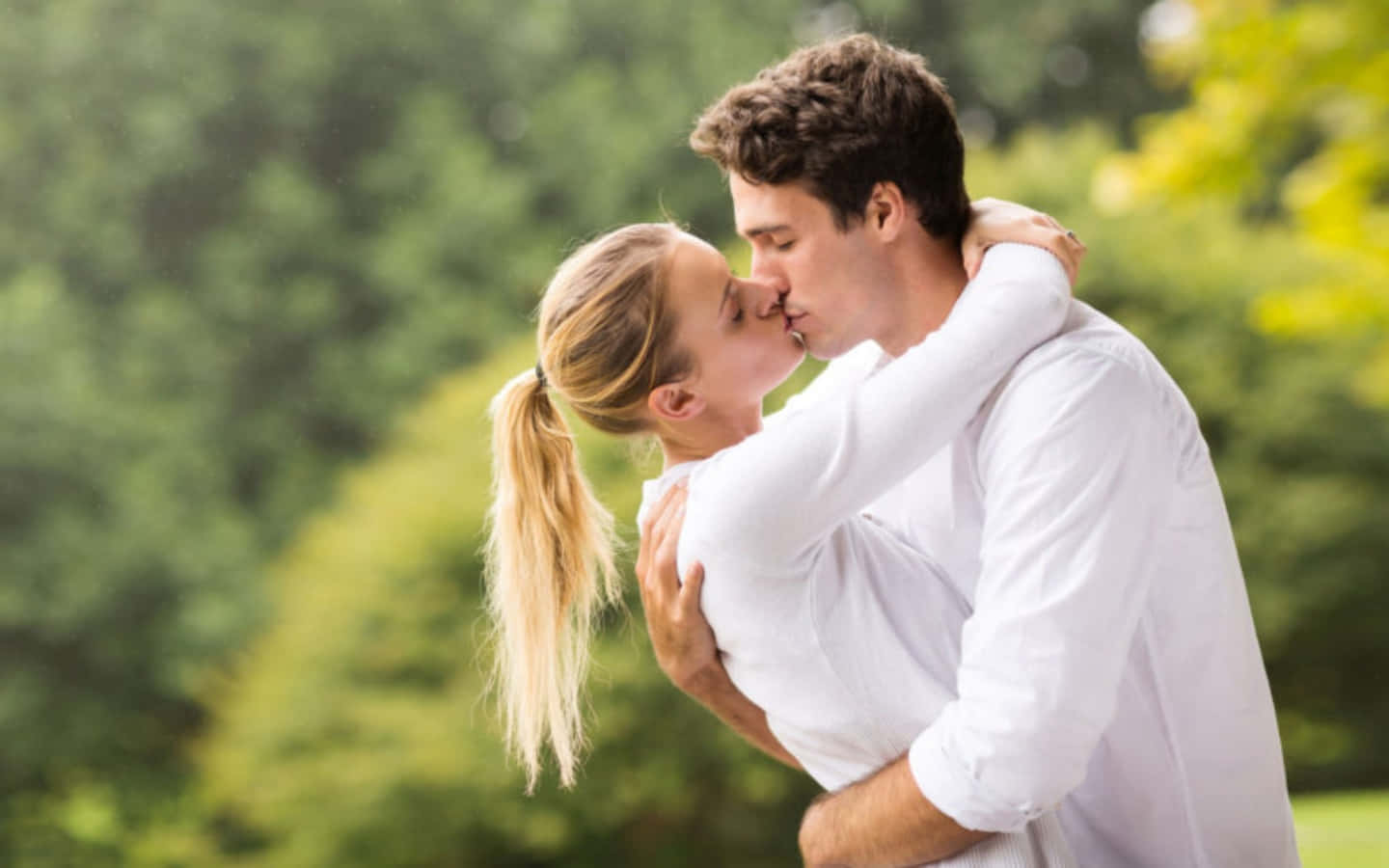 Kissing 桌布 壁紙 — 一對情侶在陽光明媚的公園裡親吻，背景中茂密的綠樹柔和地模糊。