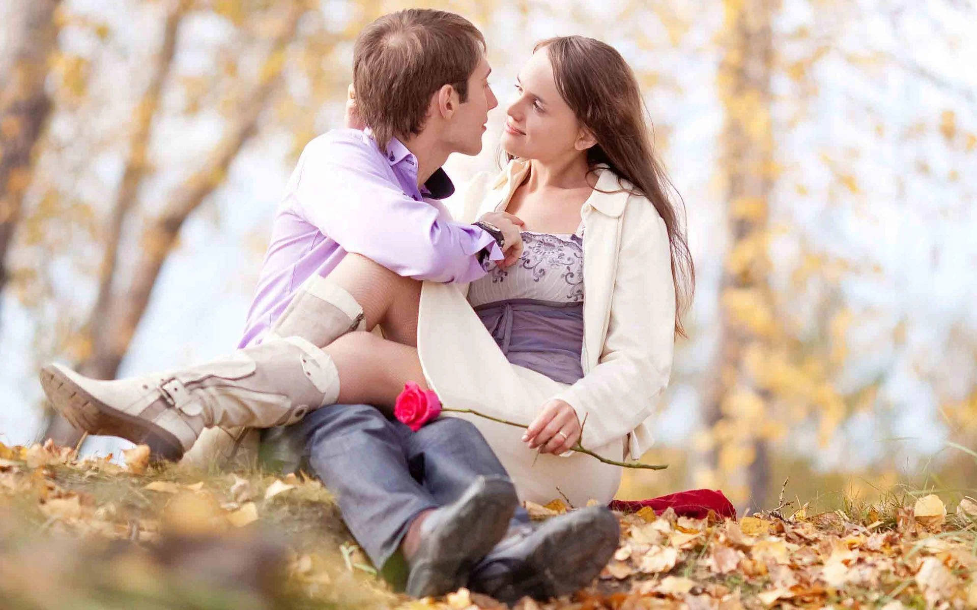 Romantic Love 桌布 壁紙 — 一對情侶在陽光明媚的秋日公園中分享親密時刻，周圍是金色的樹葉和一朵紅玫瑰。