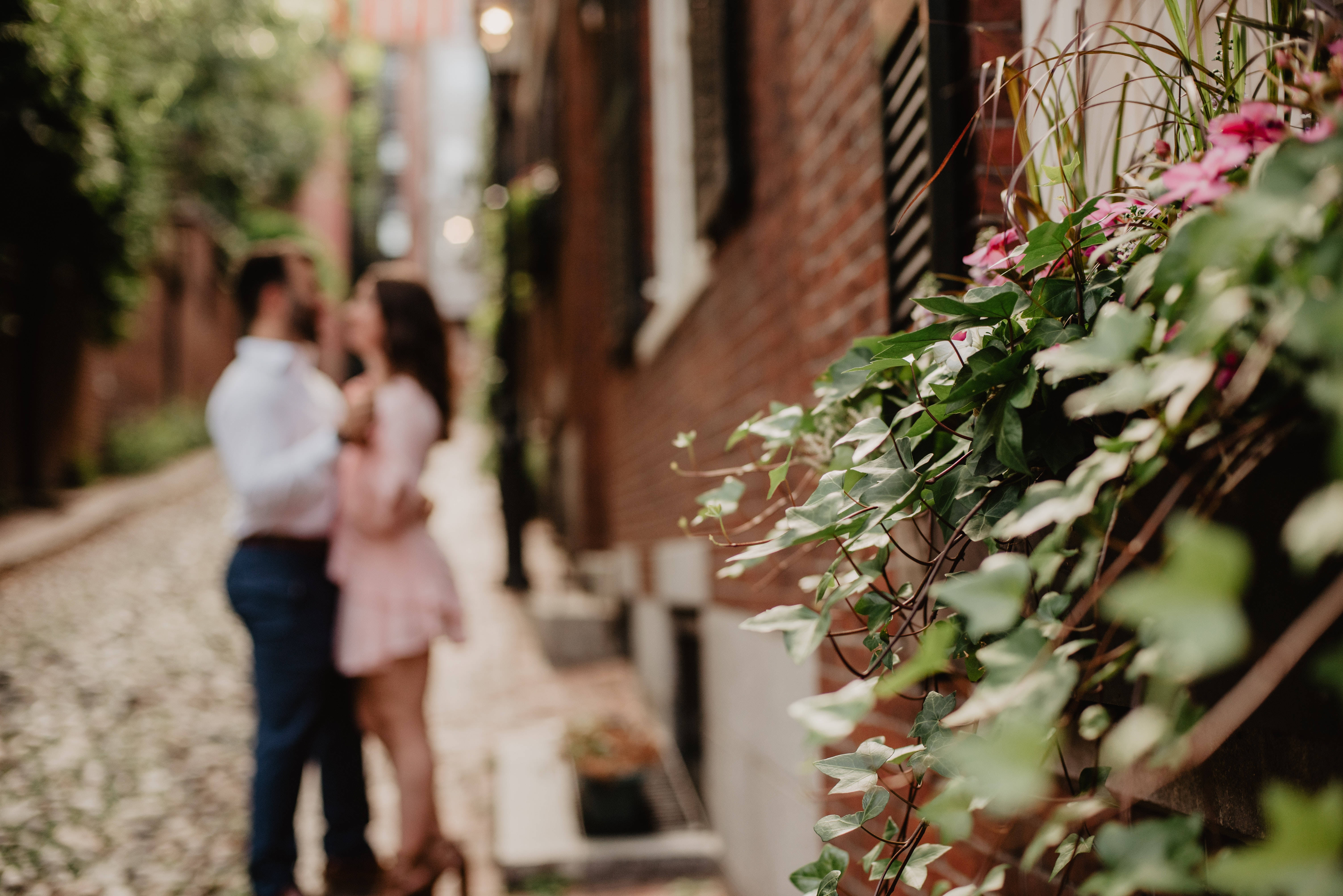 Romantic Love 桌布 壁紙 — 一對情侶在陽光灑落的磚砌小巷中溫柔相擁，四周環繞著茂密的綠植和盛開的粉紅花朵。