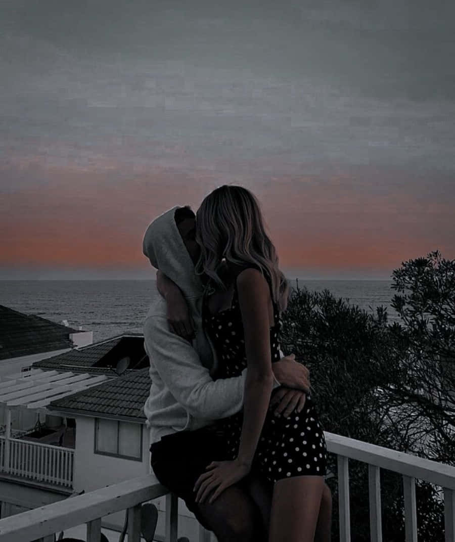 Hug 桌布 壁紙 — 一對情侶在陽台上擁抱，而憂鬱的夕陽在海洋上空以柔和的粉紅色和灰色色調塗抹天空。