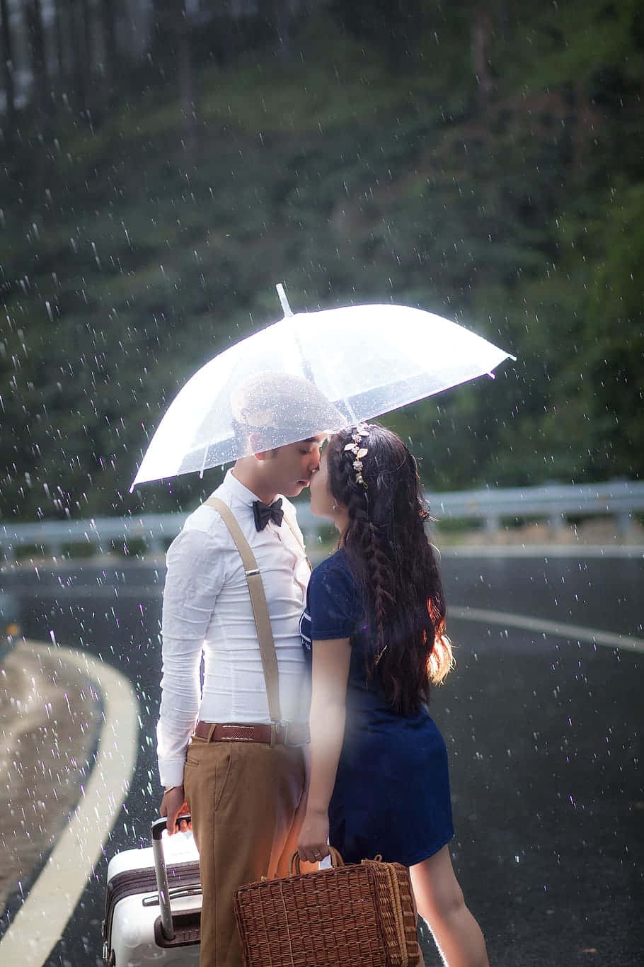 Road 桌布 壁紙 — 一對情侶在雨中的路邊共享透明雨傘和溫柔的親吻，周圍是茂密的綠樹，並手持復古行李箱。