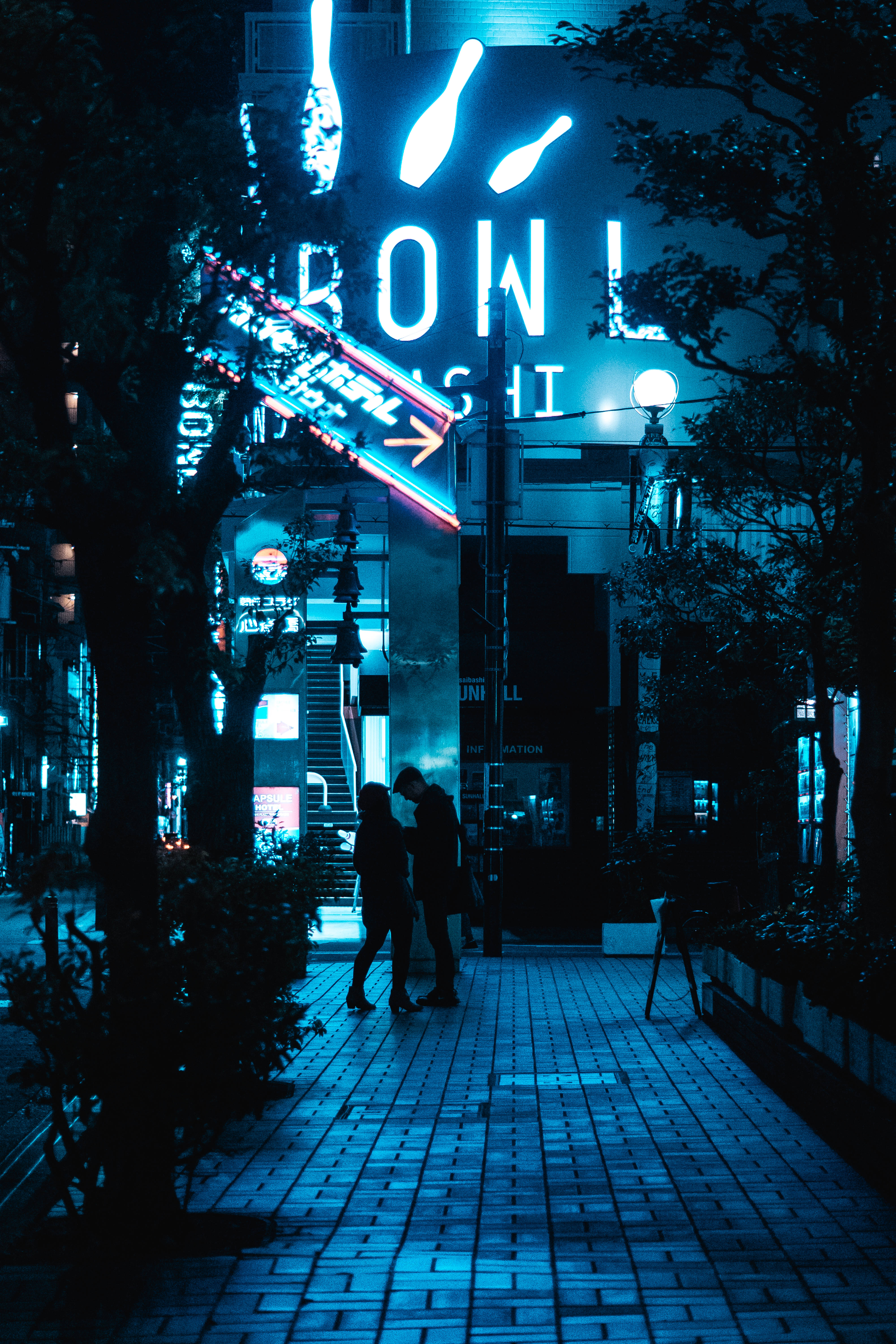 藍色 桌布 壁紙 — 一對情侶在雨夜的街道上，在發光的霓虹燈招牌下行走，街道沐浴在電藍色燈光中。