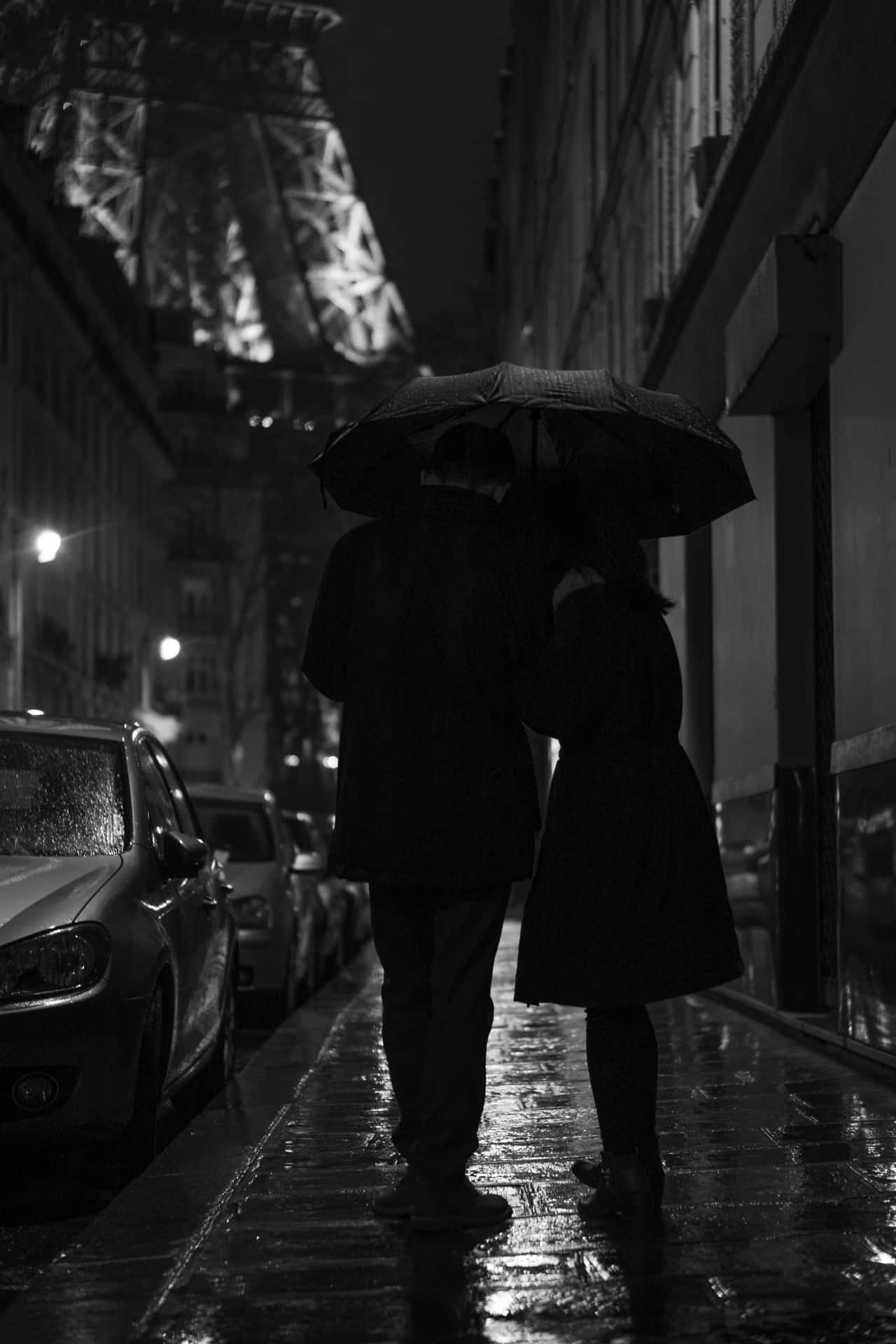 黑白 桌布 壁紙 — 一對情侶在雨濕的夜晚街道上共用一把傘，遠處的艾菲爾鐵塔閃爍著光芒。