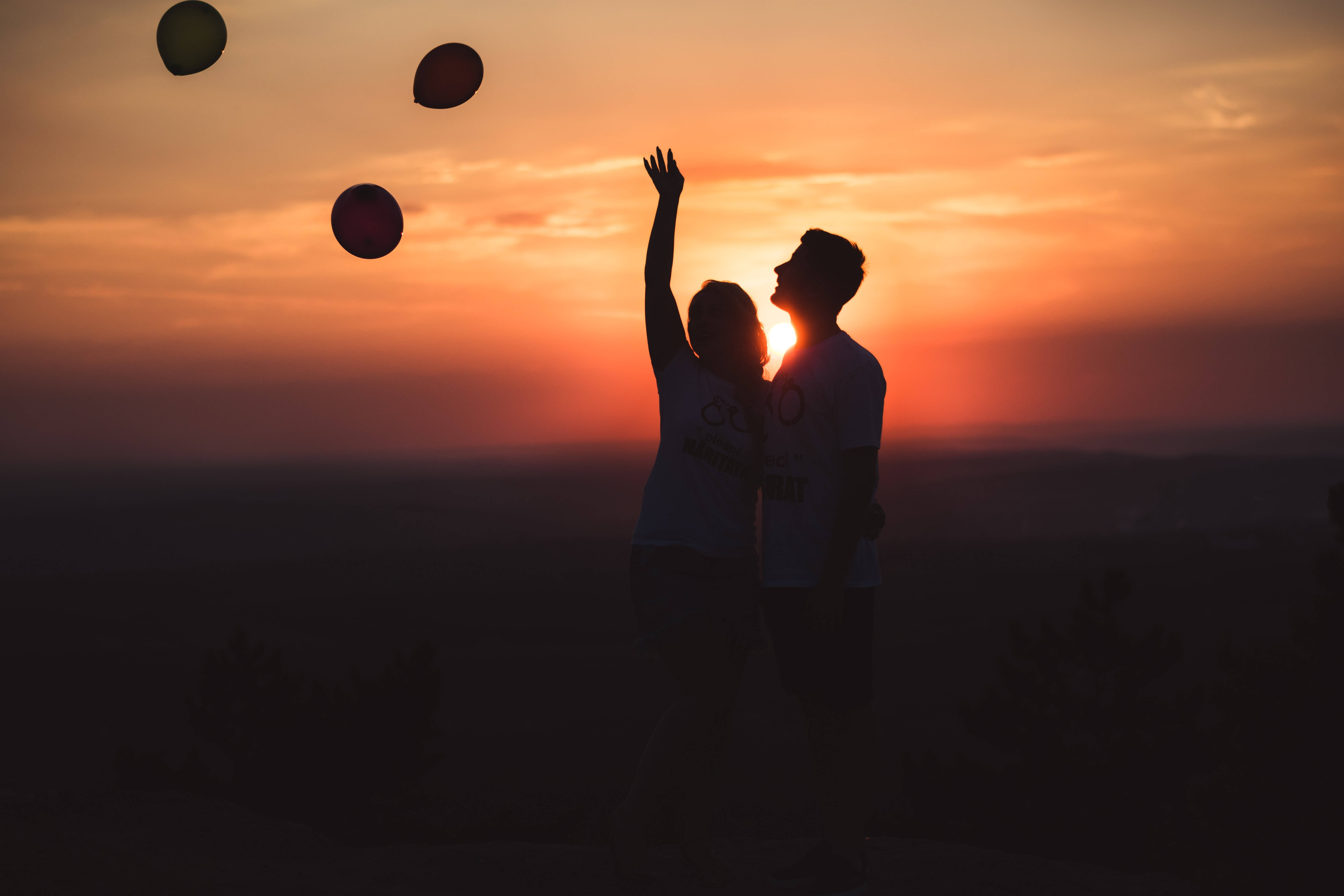 Silhouette 桌布 壁紙 — 一對情侶在鮮豔的橙色日落中形成剪影，伸手去抓漂浮在天空中的彩色氣球。