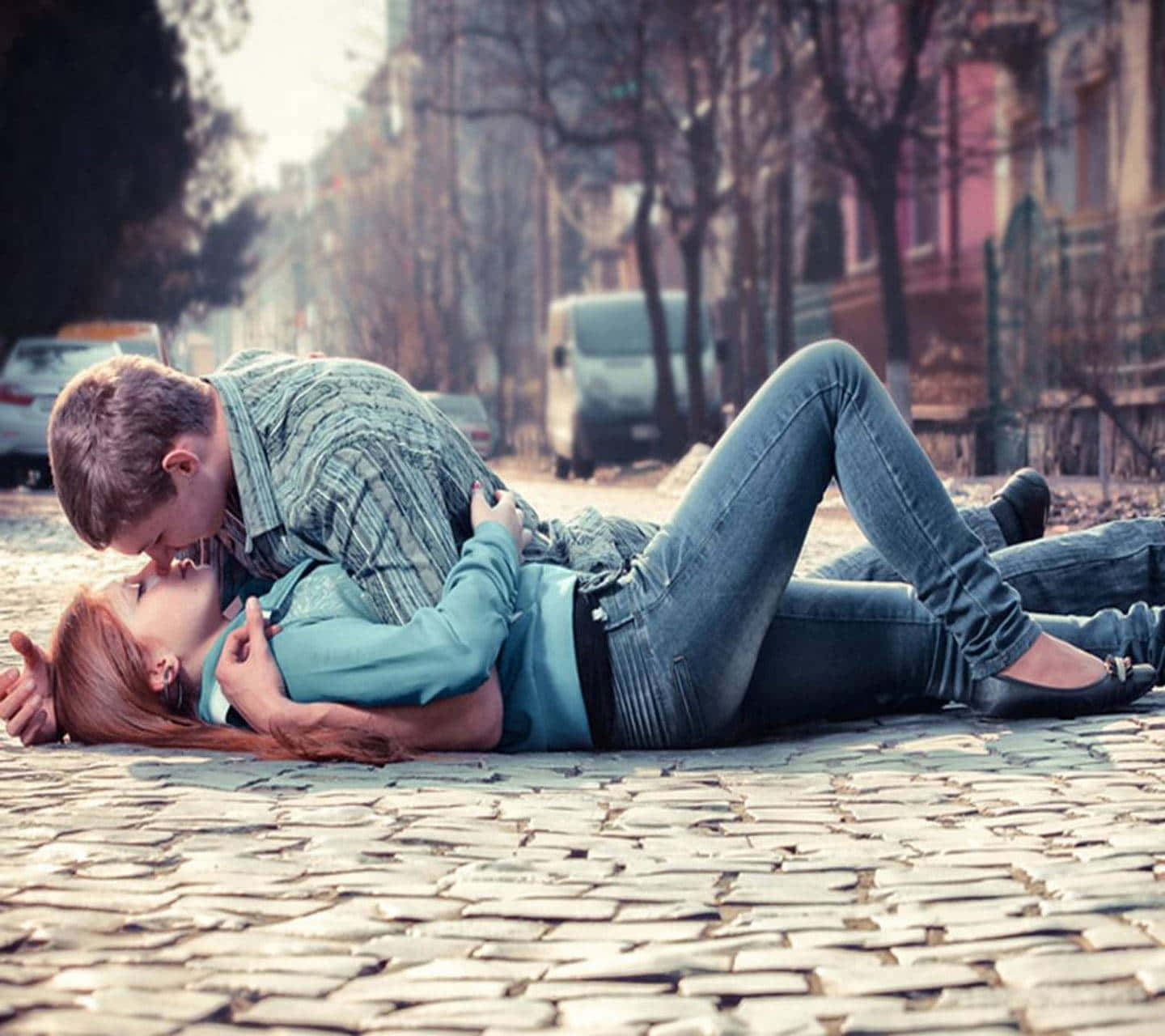 couple kiss 桌布 壁紙 — 一對情侶在鵝卵石街道上親密接吻，背景中光禿的樹木和停放的汽車模糊不清。