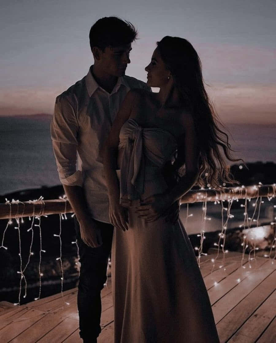 Aesthetic Couple 桌布 壁紙 — 一對情侶在黃昏時分在木甲板上擁抱，燈串在柔和的紫色和橙色天空下閃耀。