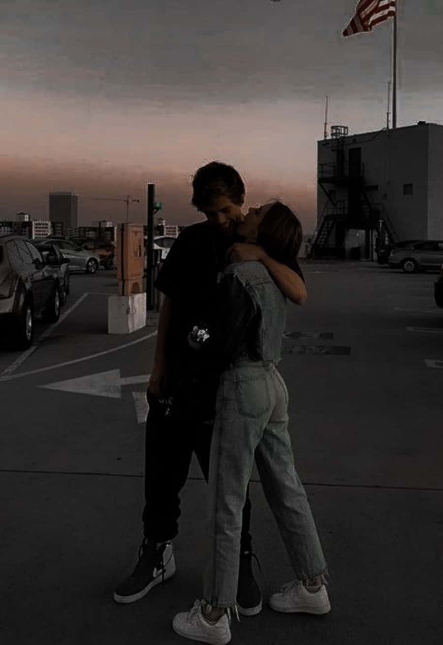 Aesthetic Couple 桌布 壁紙 — 一對情侶在黃昏時分昏暗的停車場熱情擁抱，背景是城市天際線和美國國旗。