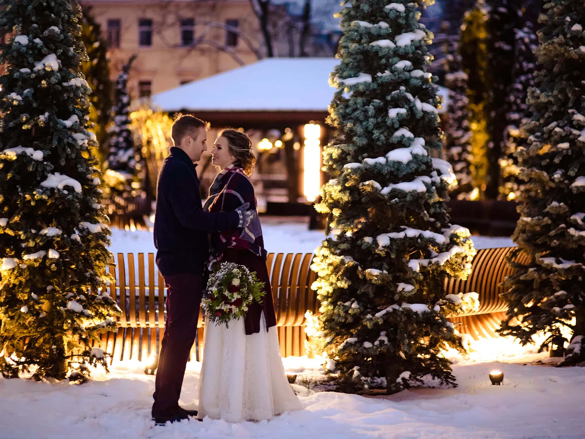 Couples Christmas 桌布 壁紙 — 一對情侶在黃昏時分的冬季公園裡，在灑滿白雪、閃爍著金色燈光的常青樹下溫暖地擁抱。