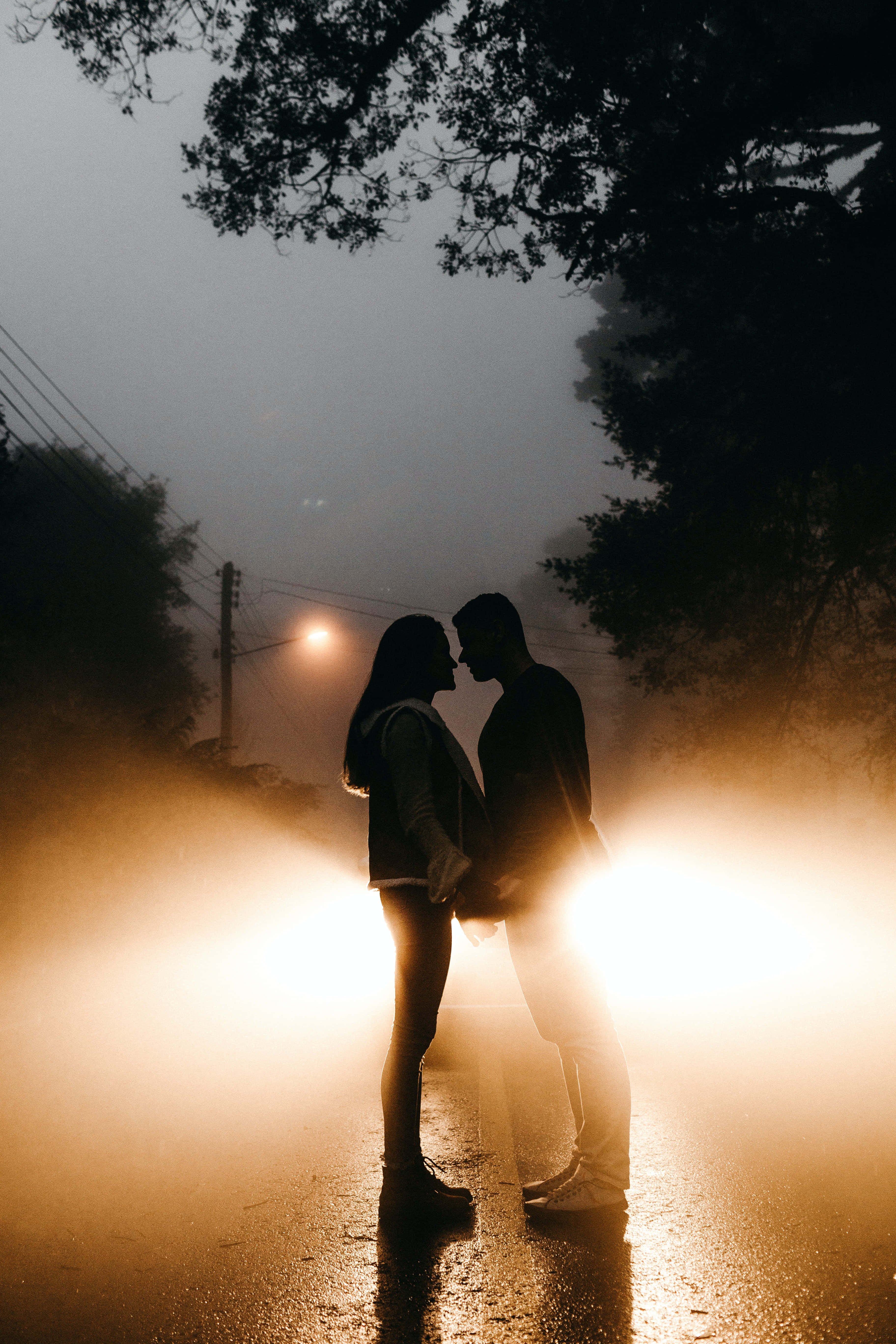 Beautiful Romantic 桌布 壁紙 — 一對情侶在黃昏時分站在霧氣瀰漫的潮濕道路上形成剪影，沐浴在車頭燈溫暖的金色光芒中。