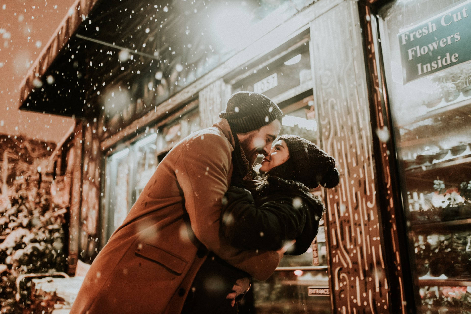 Love Story 桌布 壁紙 — 一對情侶在黃昏時分花店外飄落的雪中溫暖擁抱，柔和的金色燈光。