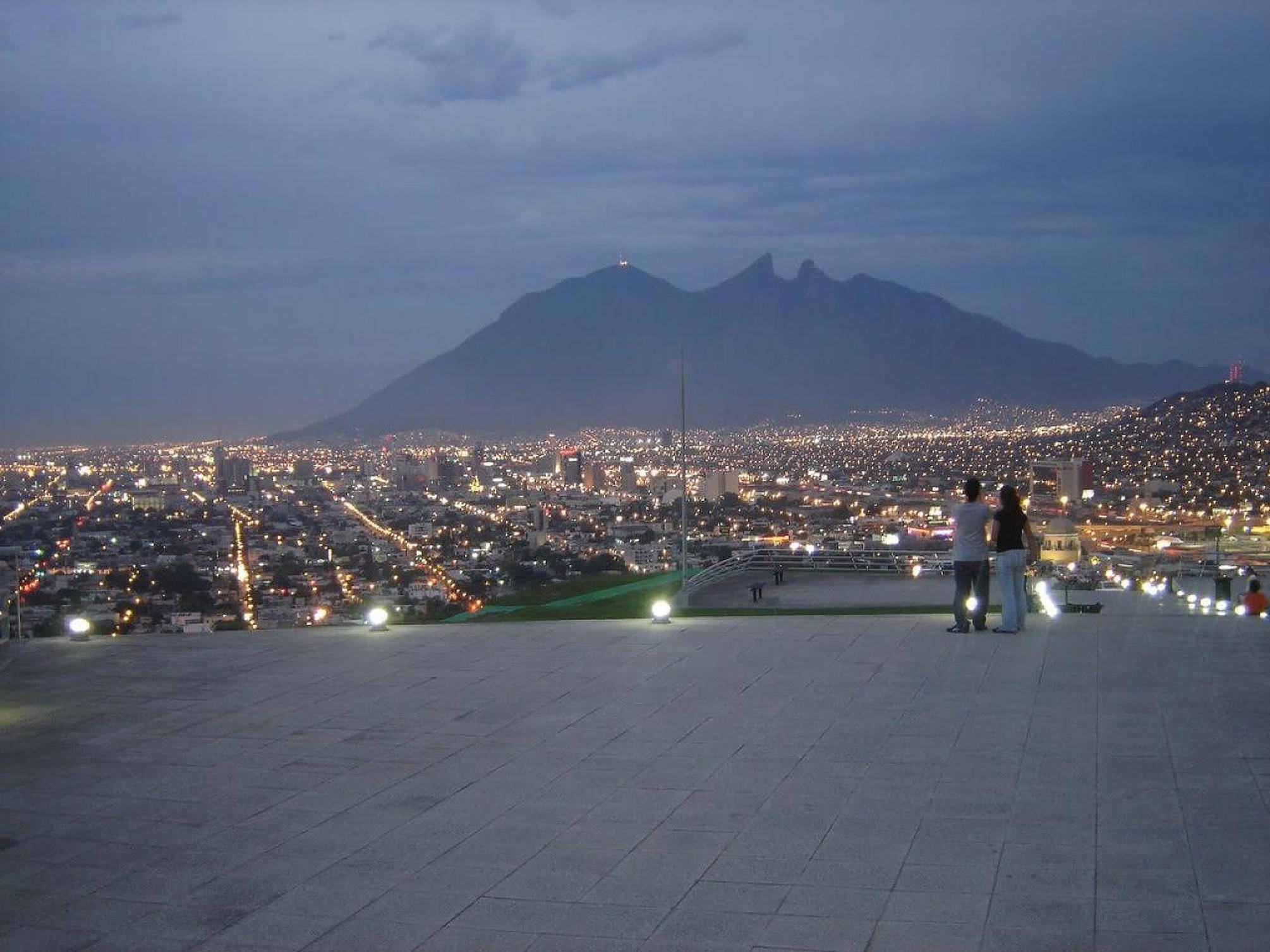 Monterrey 桌布 壁紙 — 一對情侶在黃昏時站在磁磚鋪設的觀景台上，眺望著被無數燈光照亮的廣闊城市景觀，背景中有一座黑暗的山巒聳立。