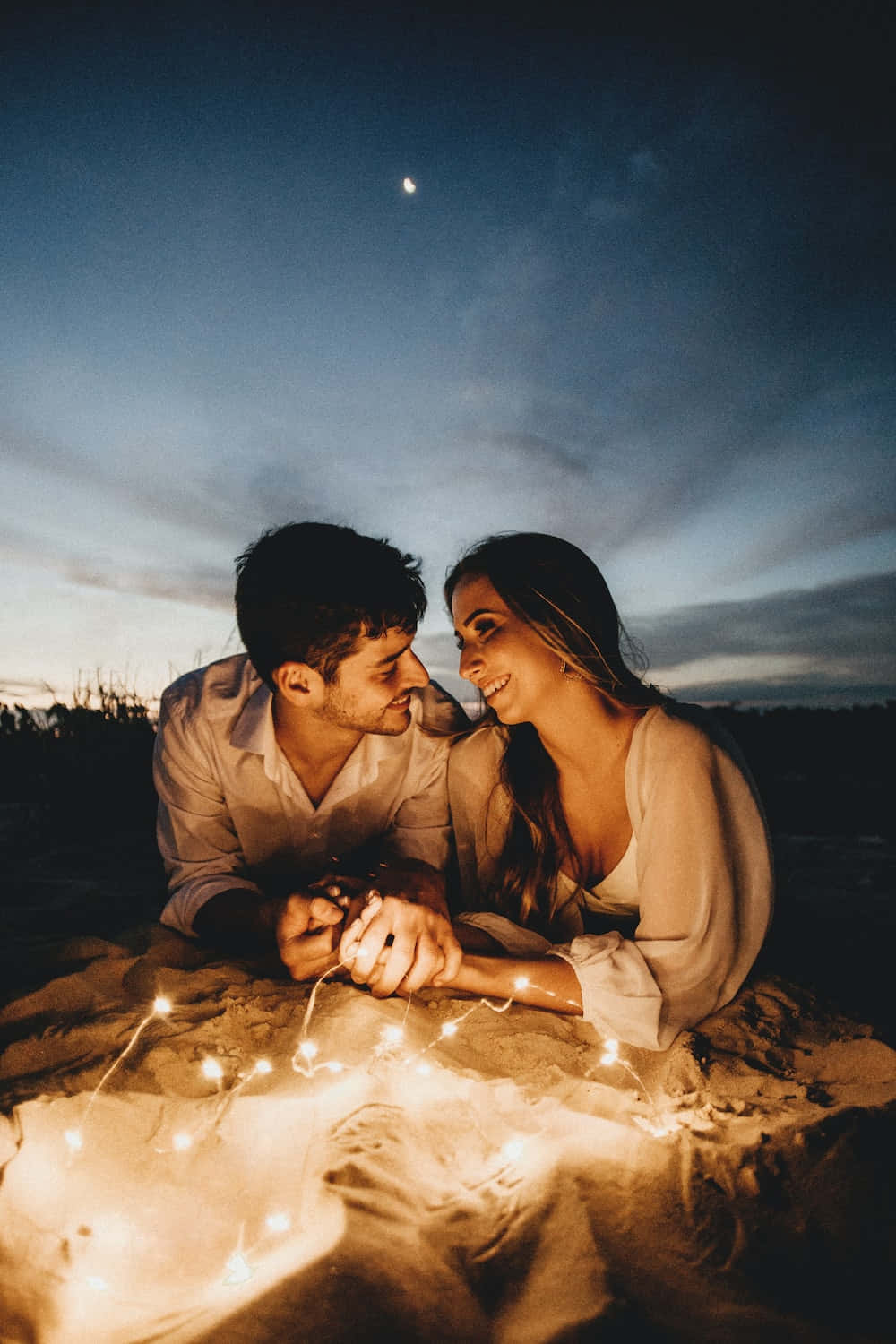 boyfriend 桌布 壁紙 — 一對情侶在黃昏的沙灘上分享溫馨時刻，被串燈和上方的彎月照亮。
