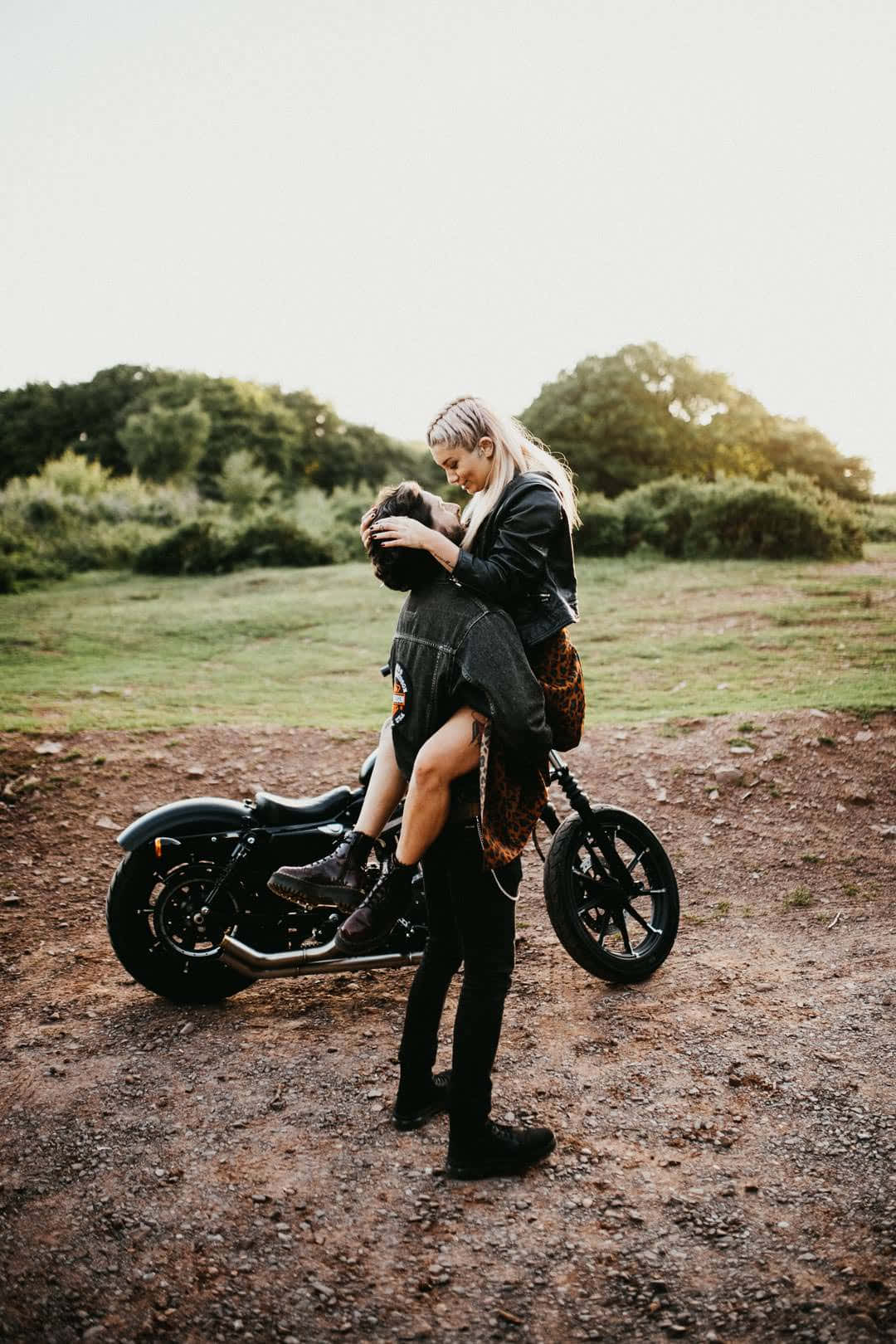 biker 桌布 壁紙 — 一對情侶在黃金時段，於一條被綠意環繞的泥濘小路上，在一輛停泊的機車旁熱情擁抱。