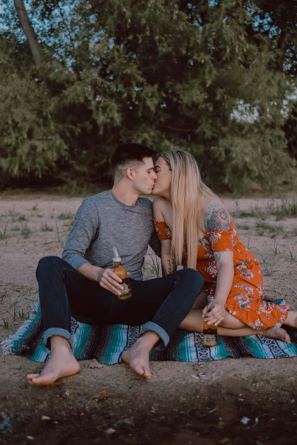 Kissing 桌布 壁紙 — 一對情侶在黃金時段於樹下色彩繽紛的毯子上熱情接吻，手中拿著啤酒瓶。