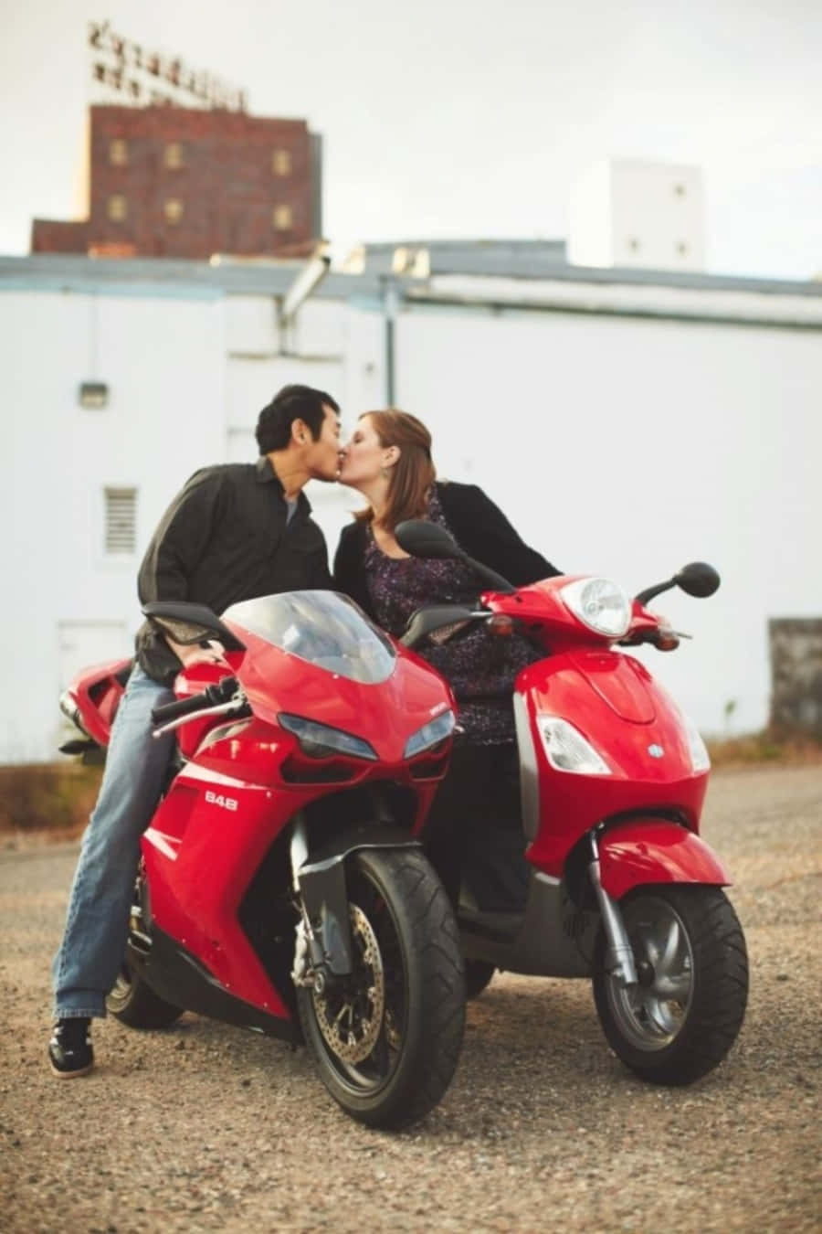 Kissing 桌布 壁紙 — 一對情侶坐在兩輛停放在工業建築附近碎石地上的紅色機車上熱情接吻。