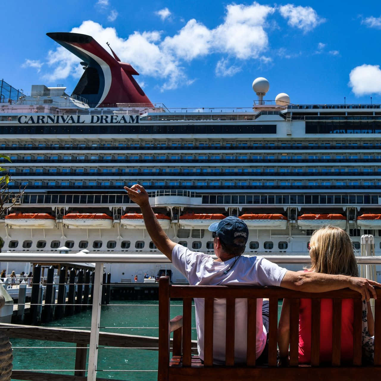 Carnival Dream 桌布 壁紙 — 一對情侶坐在長椅上，指著停泊在藍天白雲下的巨型嘉年華夢想號郵輪。