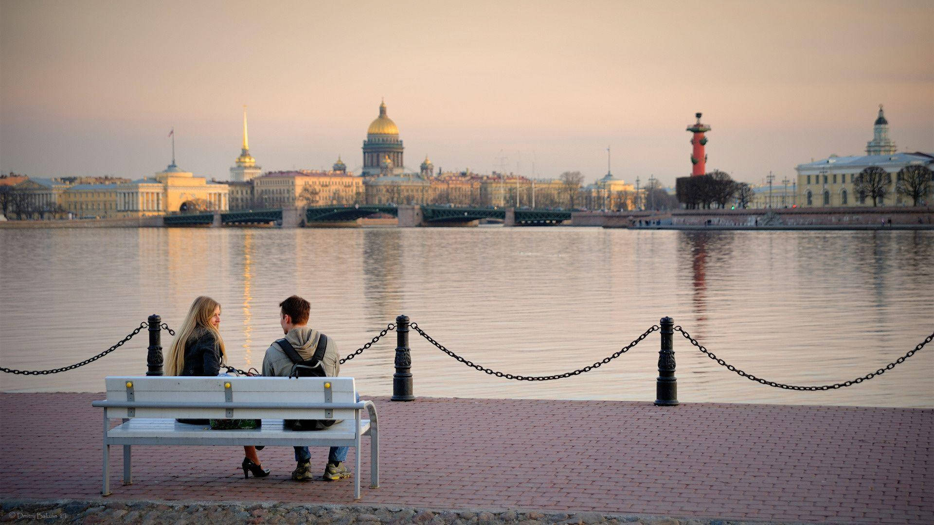 Saint Petersburg 桌布 壁紙 — 一對情侶坐在黃昏時平靜的河邊長椅上，凝視著歷史悠久的城市天際線，金色的圓頂和紅色燈塔倒映在水中。