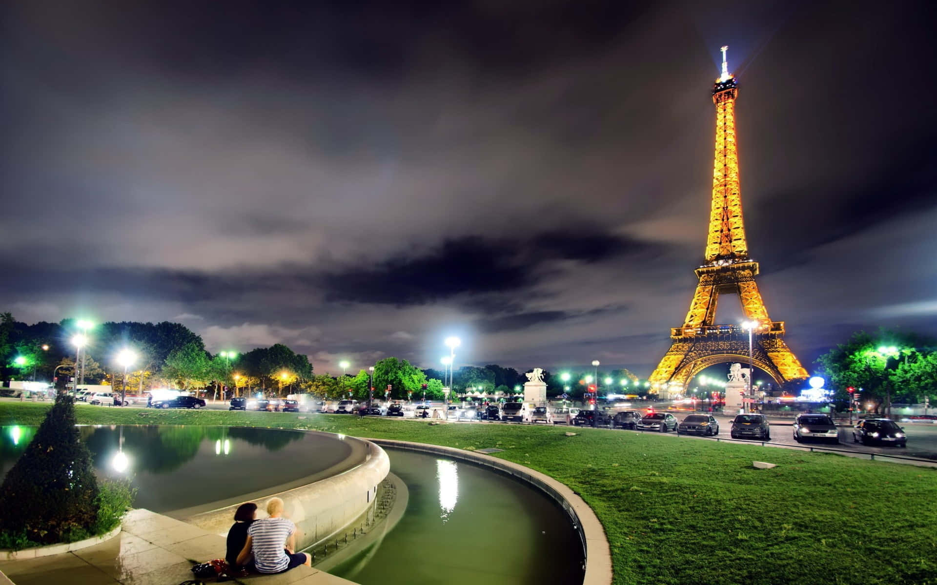 Eiffel Tower 桌布 壁紙 — 一對情侶夜晚坐在彎曲的噴泉旁，艾菲爾鐵塔在陰暗多雲的天空中閃耀著金色光芒。