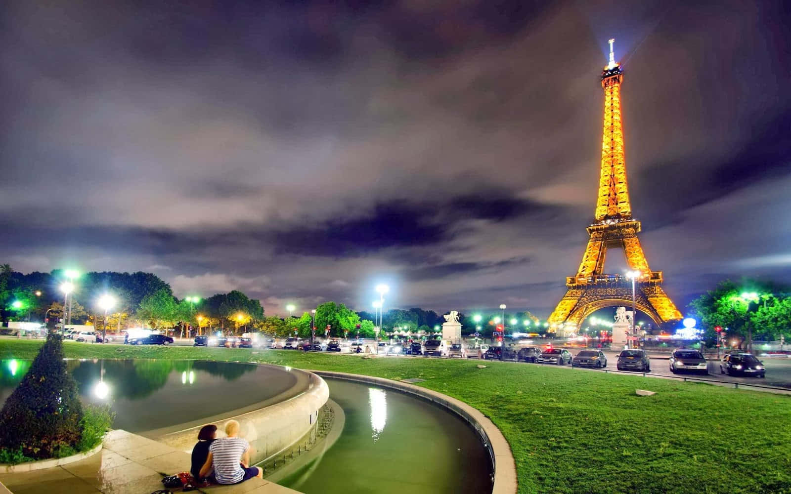 France 桌布 壁紙 — 一對情侶夜晚坐在彎曲的噴泉旁，艾菲爾鐵塔在陰雲密布的天空下閃耀著金色光芒。