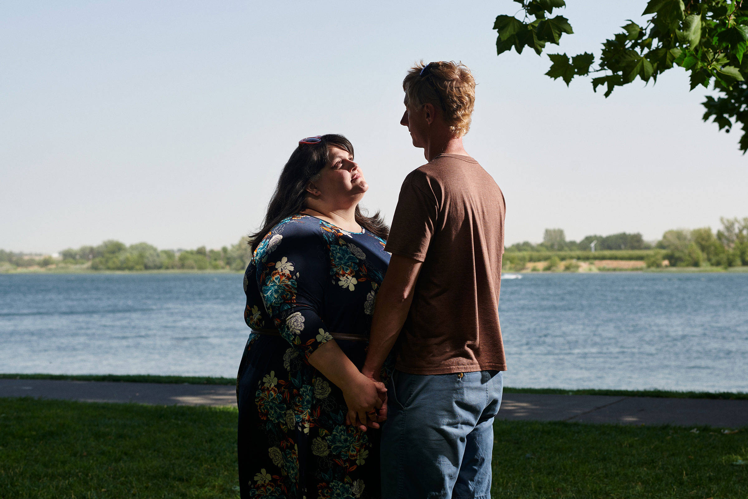 couple love 桌布 壁紙 — 一對情侶手牽手，在平靜的湖邊，明亮的天空下，綠樹環繞的場景中互相凝視。