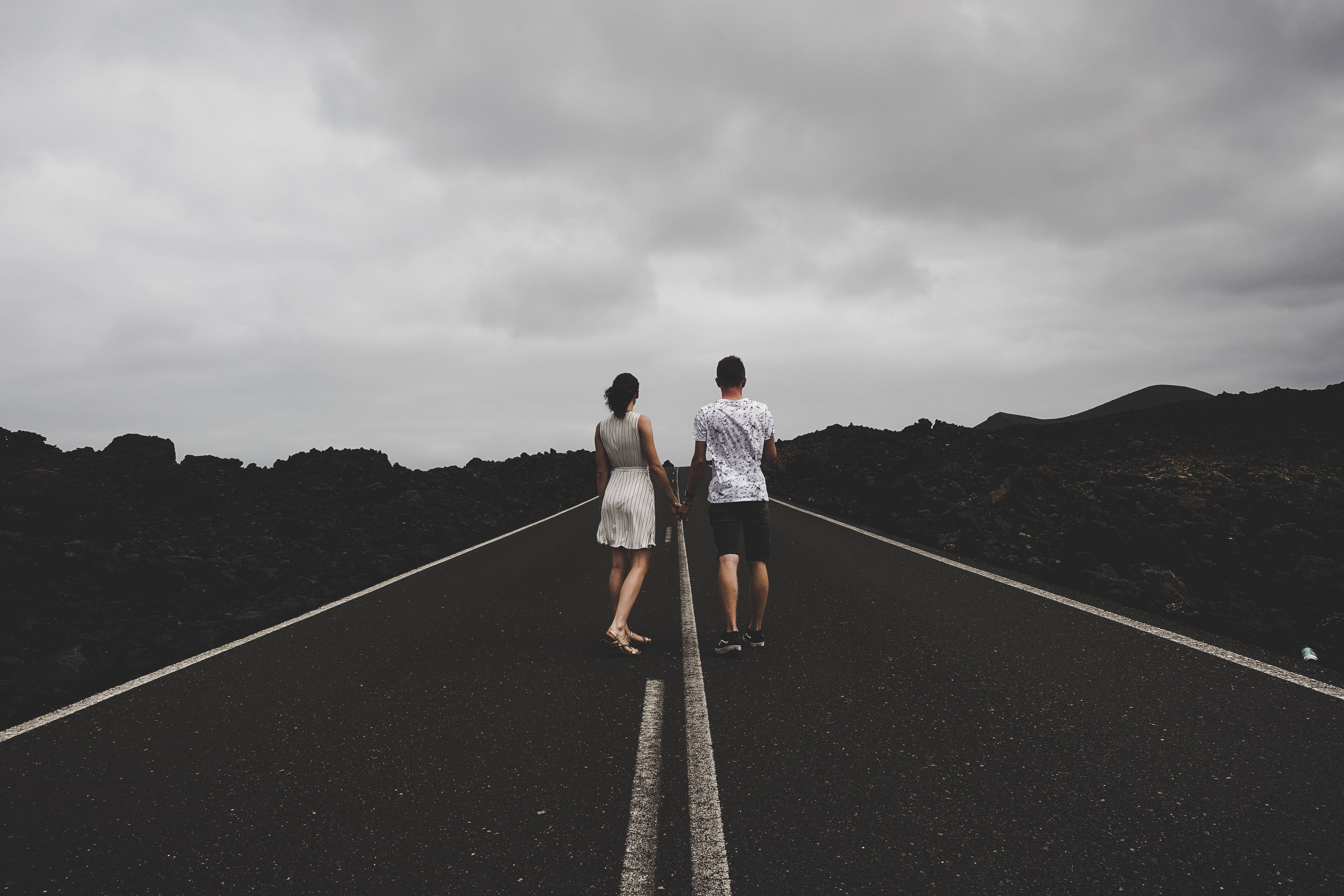 Road 桌布 壁紙 — 一對情侶手牽手，沿著一條長長的空蕩蕩的道路行走，兩旁是黑色的火山岩，天空多雲。