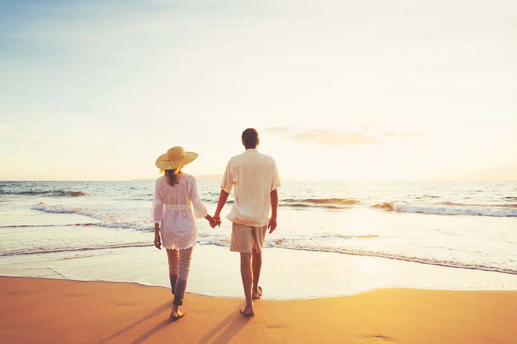 Romance 桌布 壁紙 — 一對情侶手牽手，沿著陽光照耀的海灘漫步，溫柔的海浪在日落時分徐徐湧來。