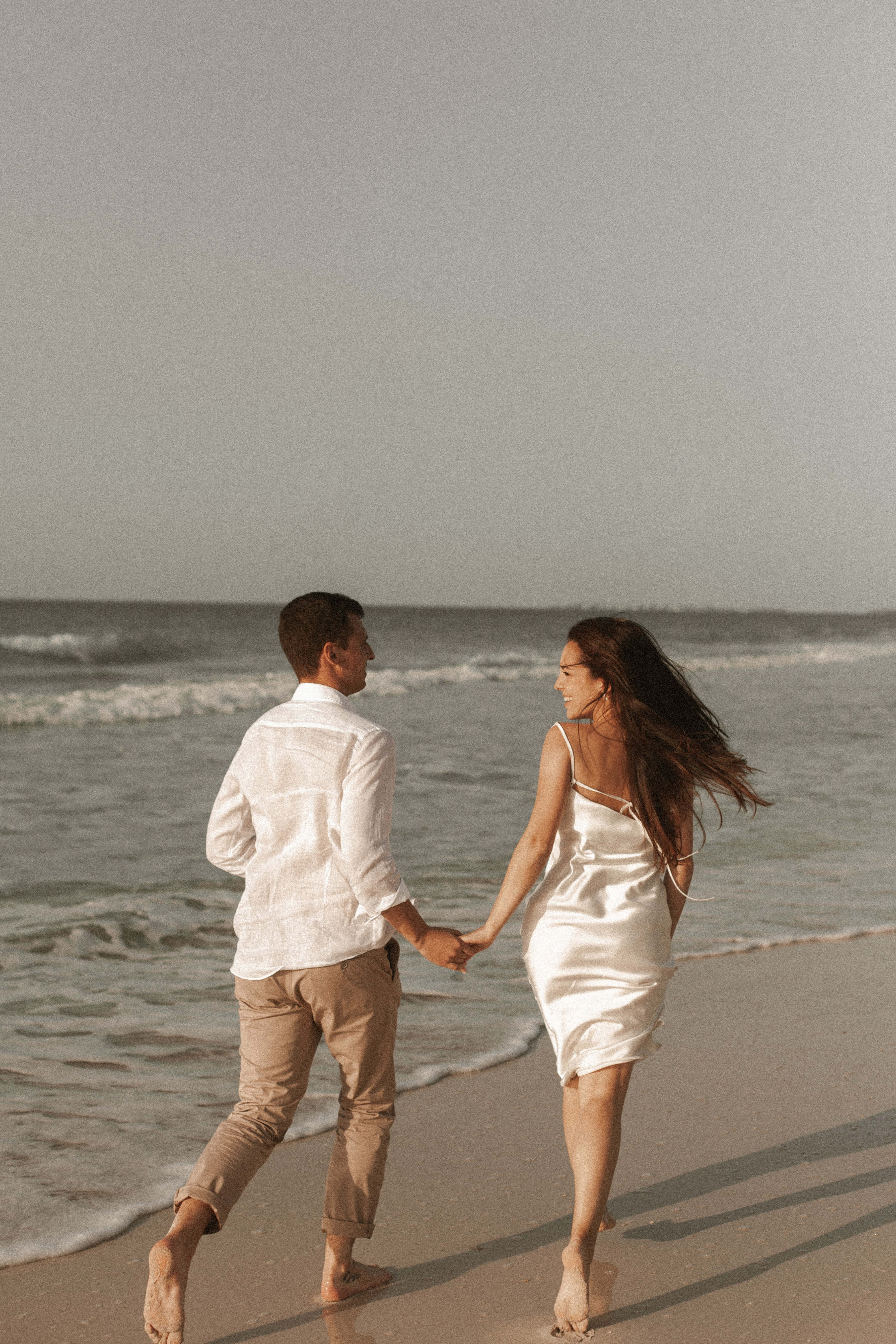 Holding Hands 桌布 壁紙 — 一對情侶手牽手，赤腳走在沙灘上，海浪在柔和的灰色天空下翻滾。