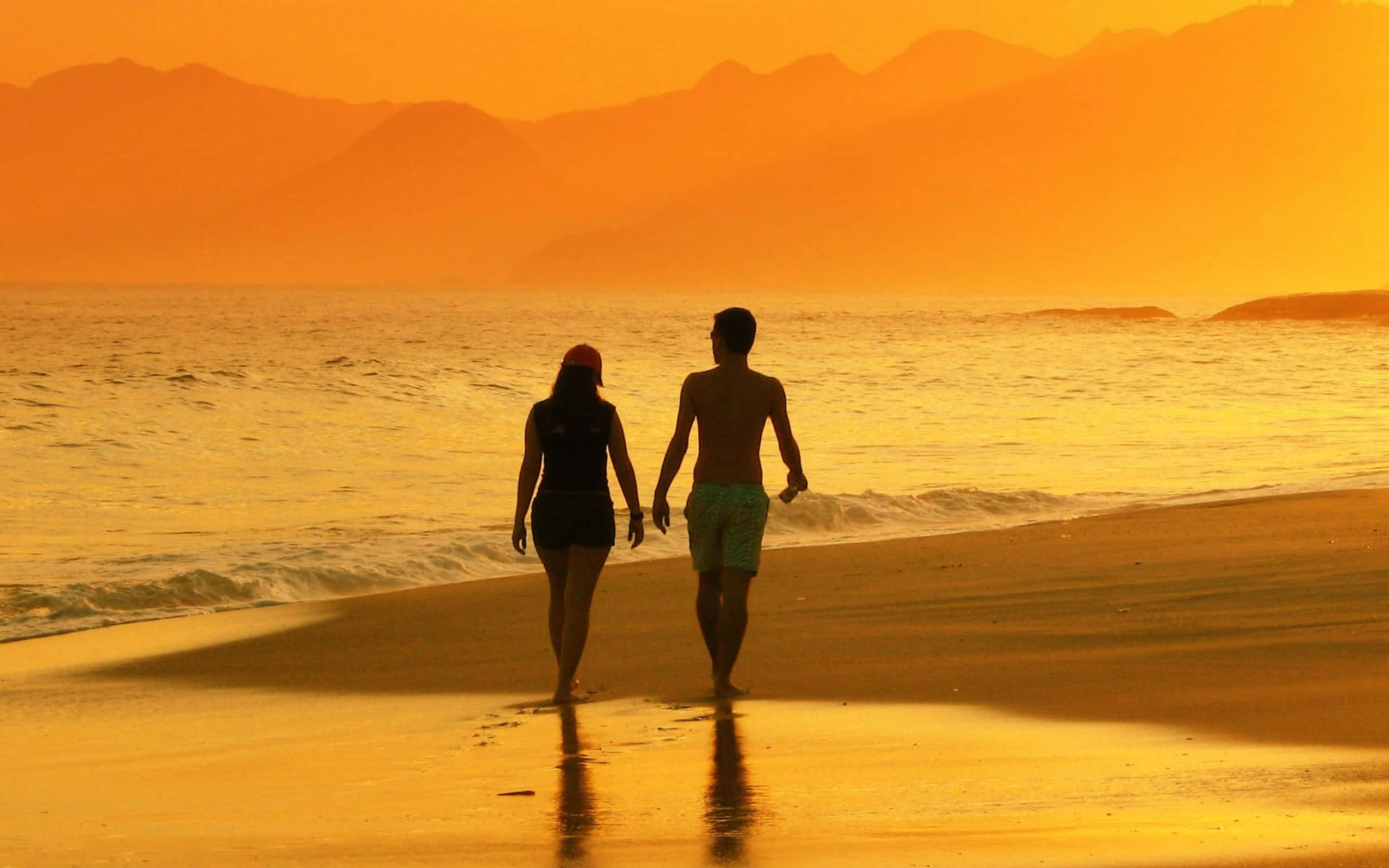 Beach Lovers 桌布 壁紙 — 一對情侶手牽手在日落時分沿著金色沙灘漫步，背景是剪影般的山巒。