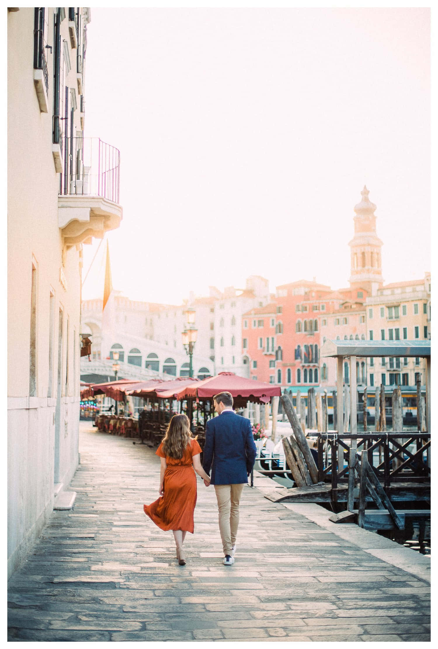 Italy Aesthetic 桌布 壁紙 — 一對情侶手牽手沿著威尼斯運河旁的小路漫步，溫暖的金色光芒灑在歷史建築和紅色雨傘上。