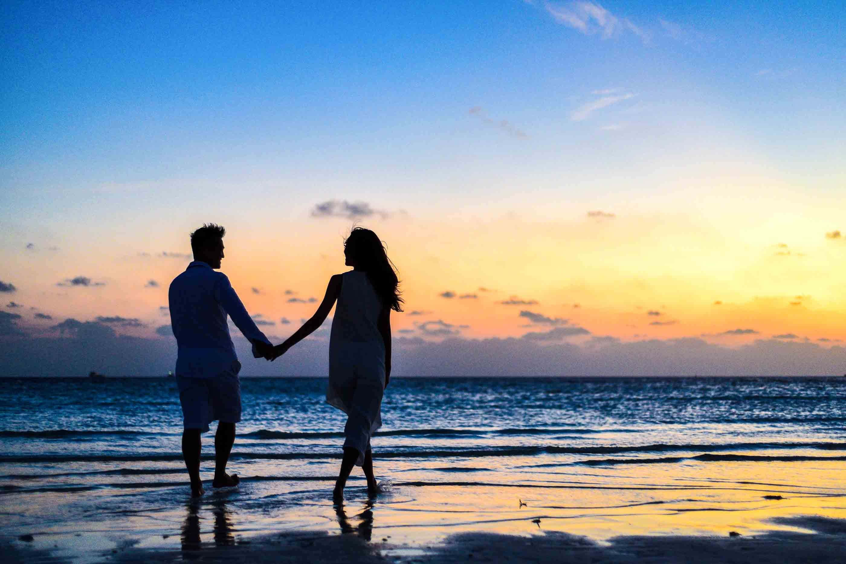 Relationship 桌布 壁紙 — 一對情侶手牽手沿著濕漉漉的海灘在日落時分漫步，他們的剪影在漸變天空下的水中倒映。