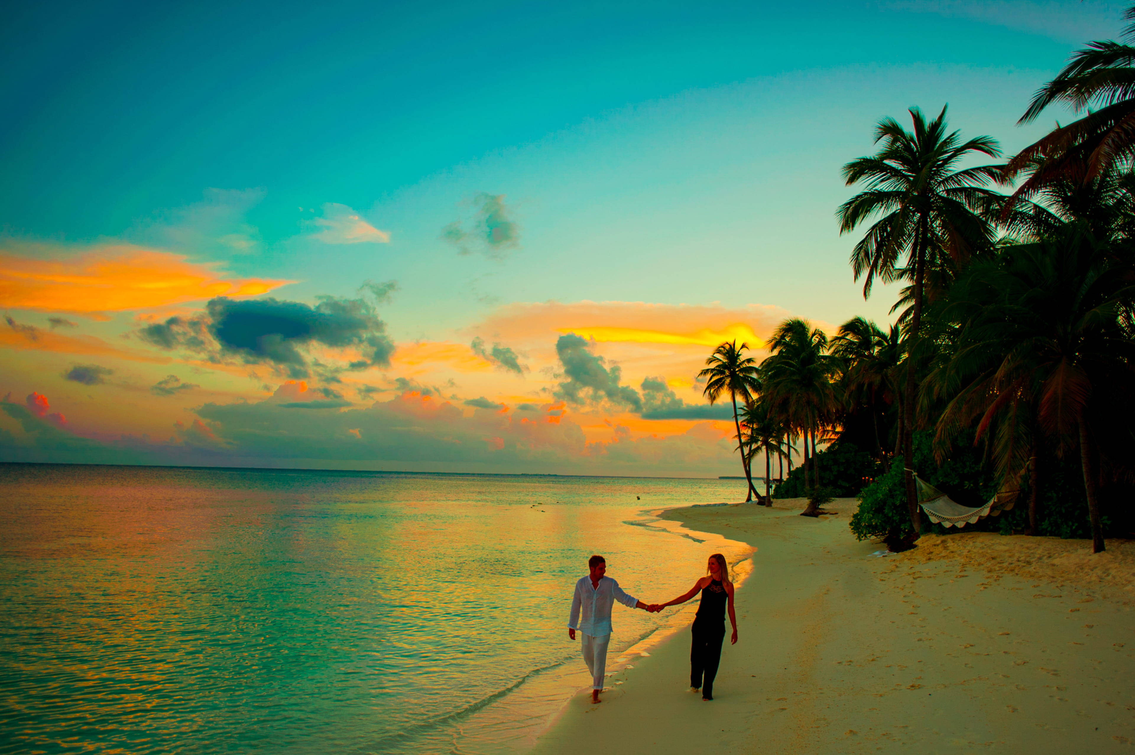 Couple 桌布 壁紙 — 一對情侶手牽手沿著熱帶海灘漫步，夕陽西下，棕櫚樹和充滿活力的橙藍色天空。