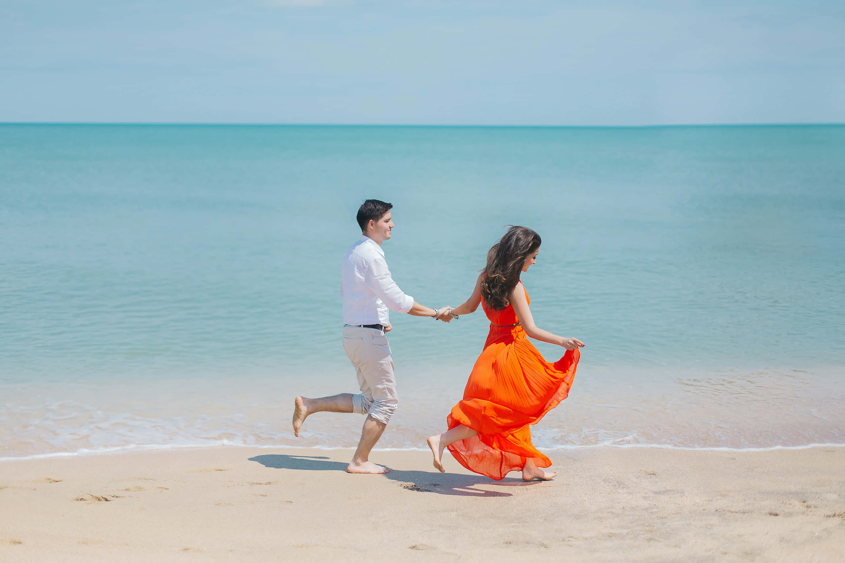 Beautiful Romantic 桌布 壁紙 — 一對情侶手牽手沿著陽光明媚的海灘奔跑，碧綠的海浪輕輕拍打著金色的沙灘。