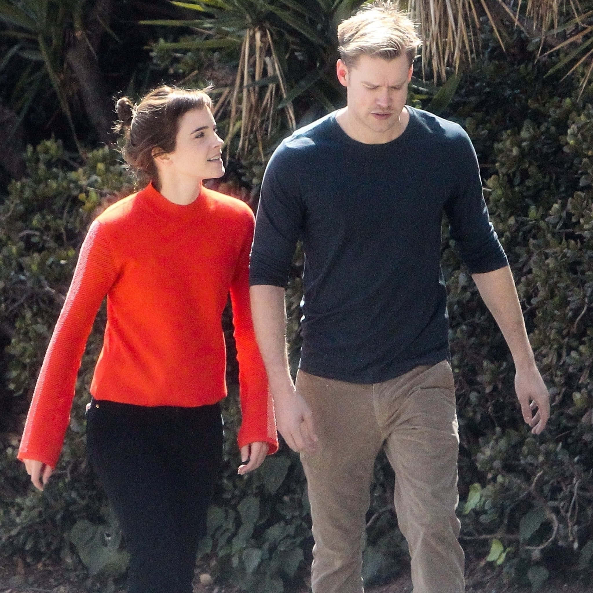 Red Sweater 桌布 壁紙 — 一對情侶手牽手穿過茂密的綠植，女士身穿亮橙色毛衣，男士身穿深色襯衫。