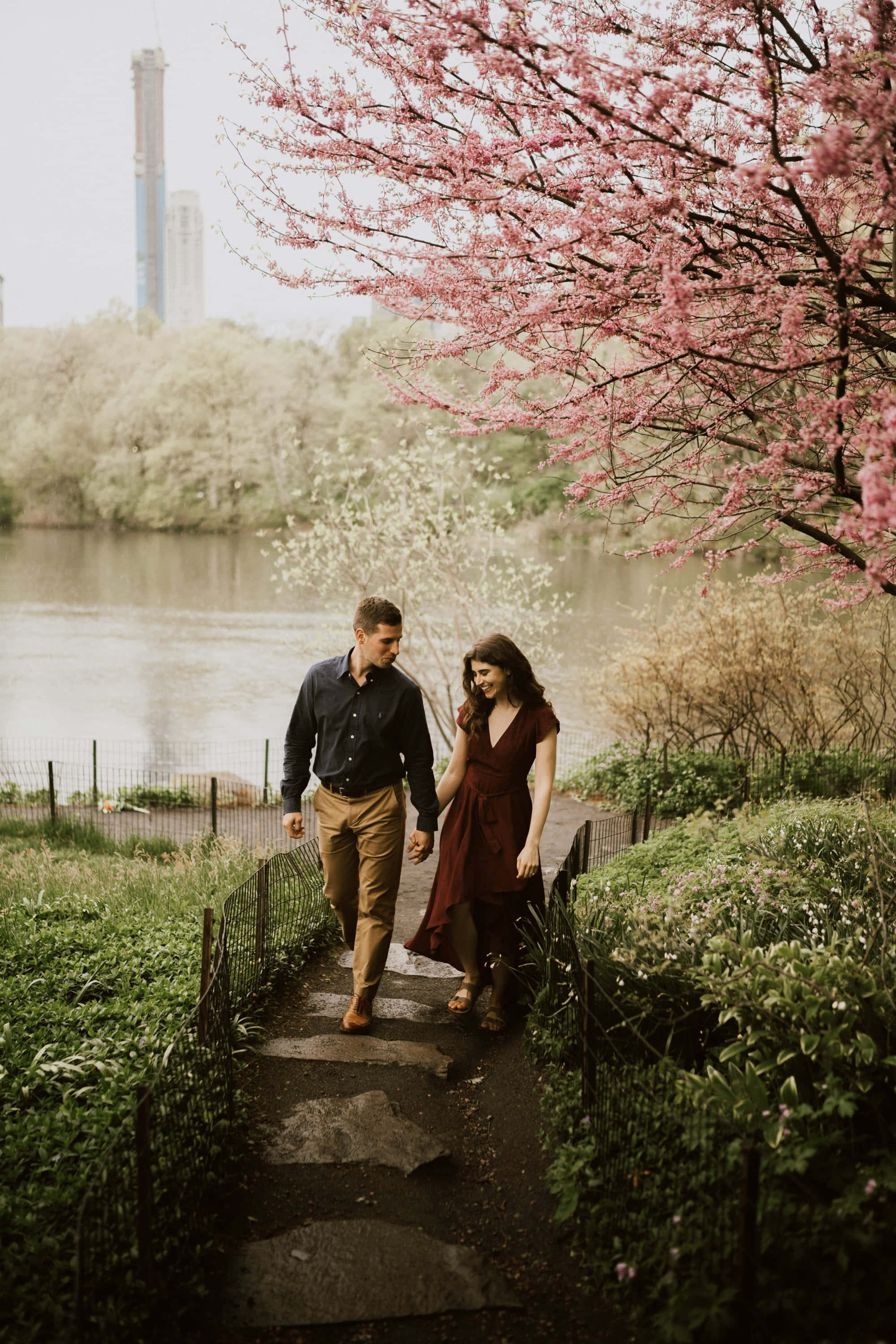Couples 桌布 壁紙 — 一對情侶手牽手走在石徑上，兩旁盛開的粉紅櫻花樹下，旁邊是平靜的湖泊，遠處隱約可見城市高樓大廈。