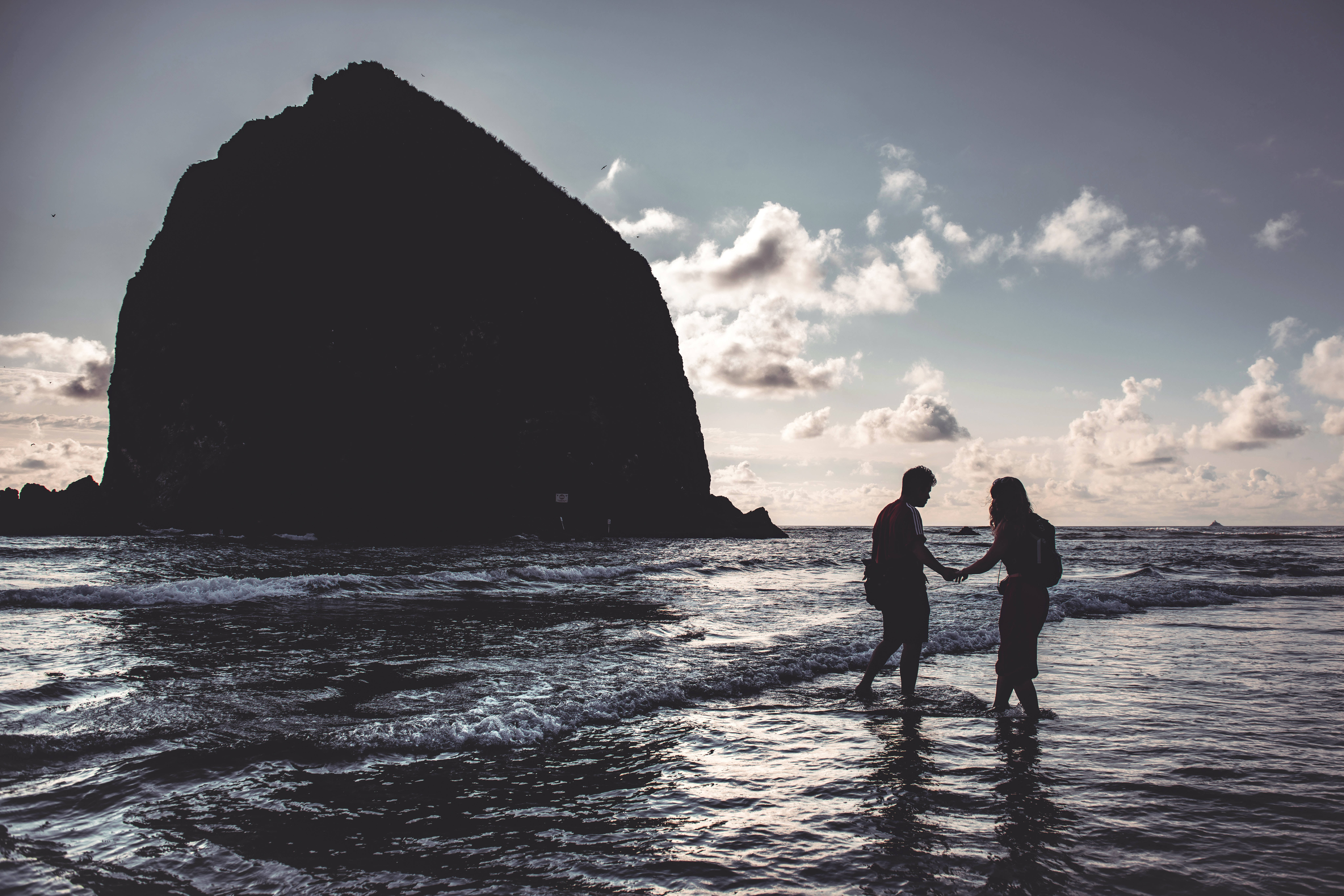 Lovers 桌布 壁紙 — 一對情侶手牽手走過淺海波浪，身後巨大的黑色岩石在陰雲密布的天空下若隱若現。