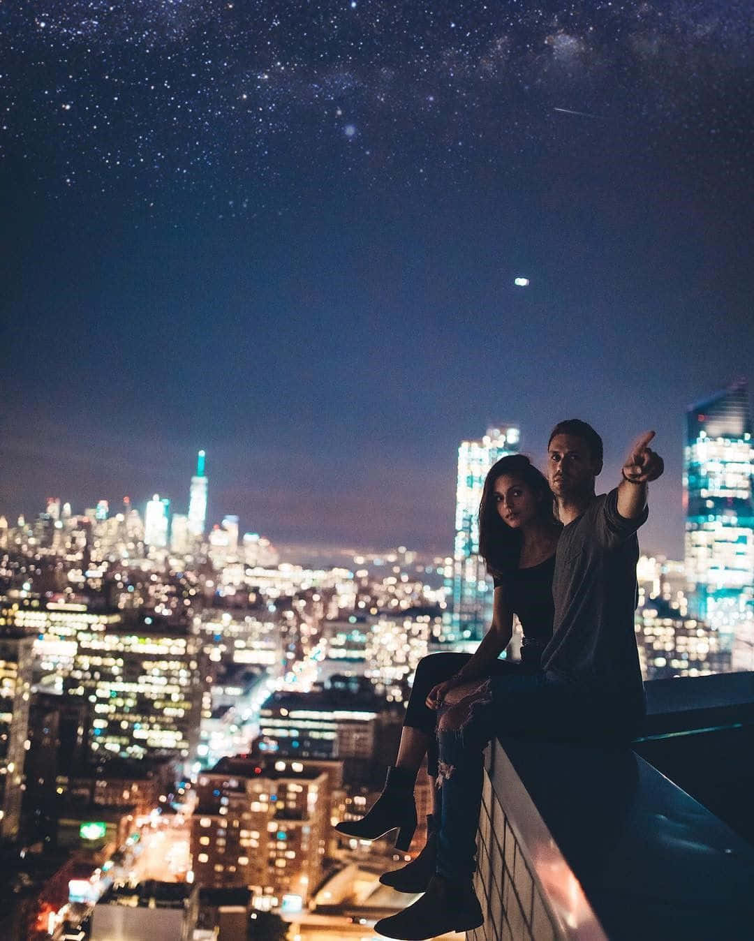 Boyfriend And Girlfriend 桌布 壁紙 — 一對情侶晚上坐在屋頂上，指著發光城市景觀上方的星空。