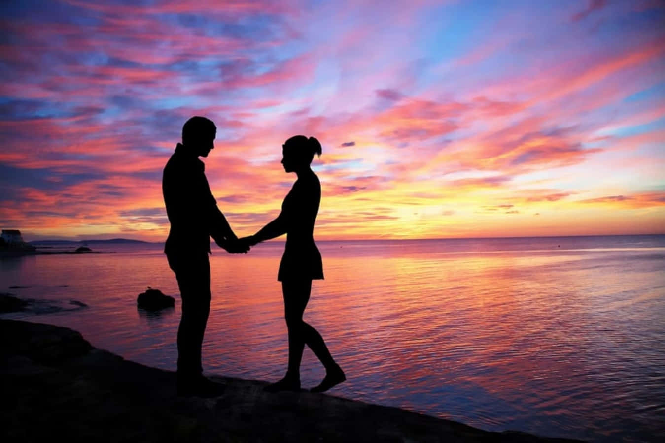 Falling In Love 桌布 壁紙 — 一對情侶的剪影在岩石海岸上牽手，鮮豔的粉紅、橙色和紫色夕陽染紅了天空和水面。