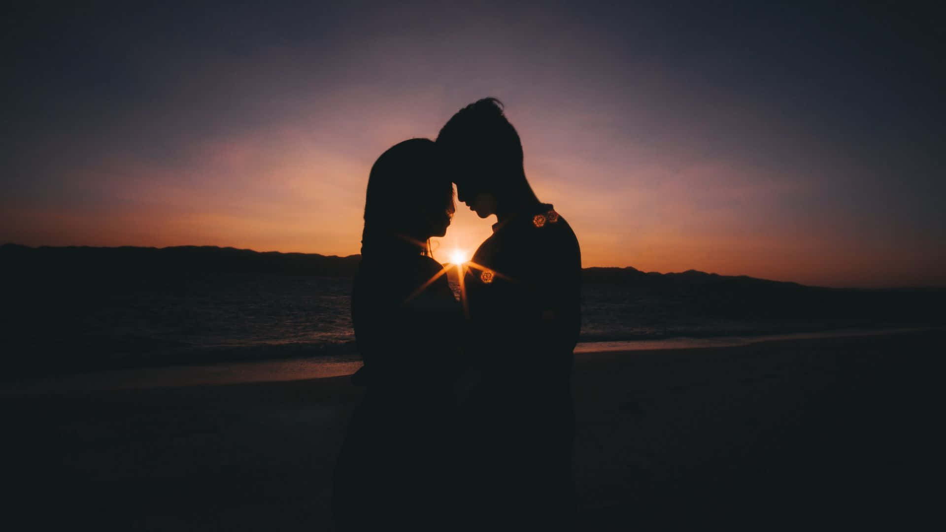 Silhouette 桌布 壁紙 — 一對情侶的剪影在海灘上擁抱，夕陽在他們身後落下，散發出溫暖的光芒。