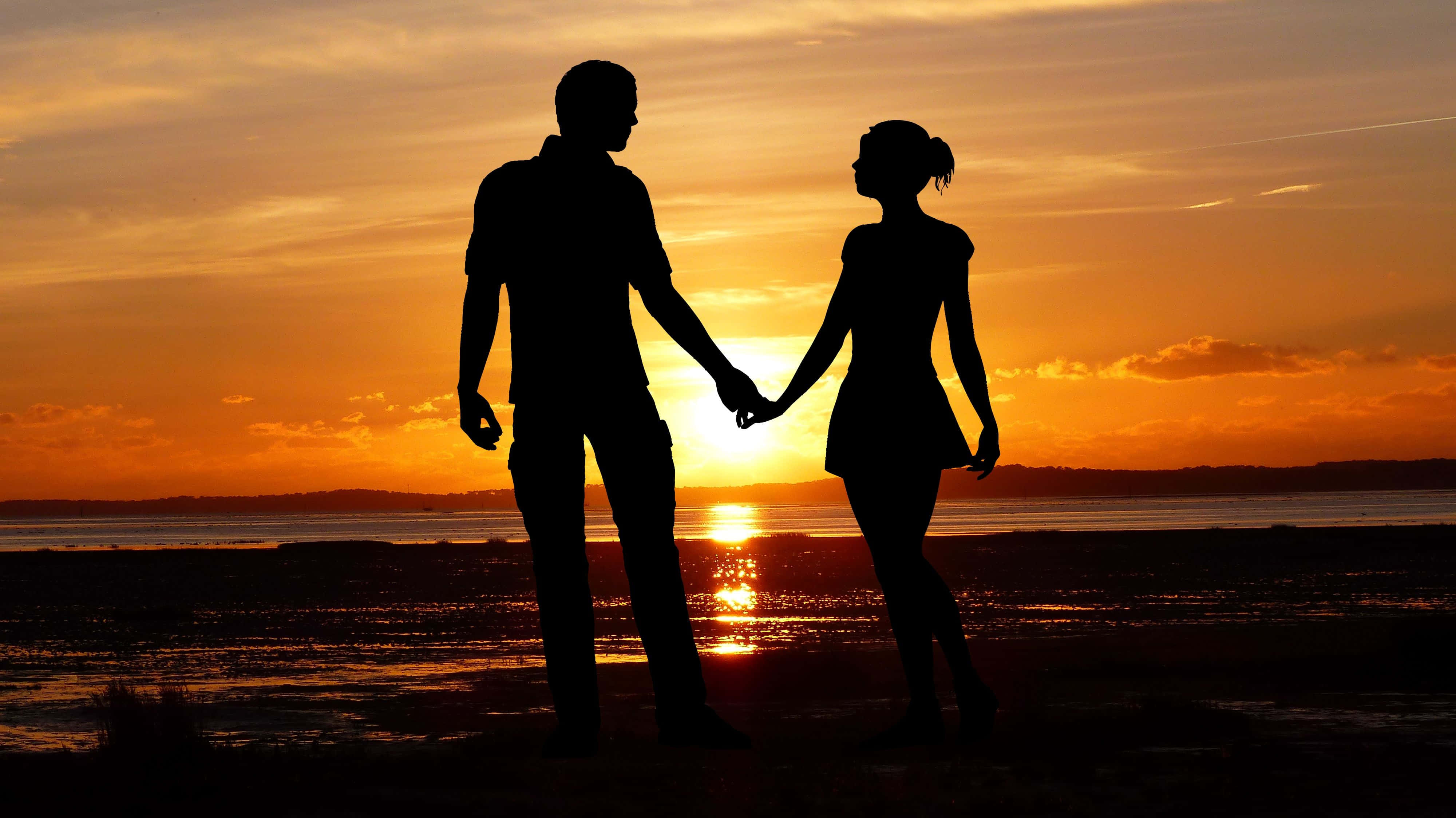 Beach Lovers 桌布 壁紙 — 一對情侶的剪影在海灘上牽手，夕陽呈現火熱的橙色和金色色調。