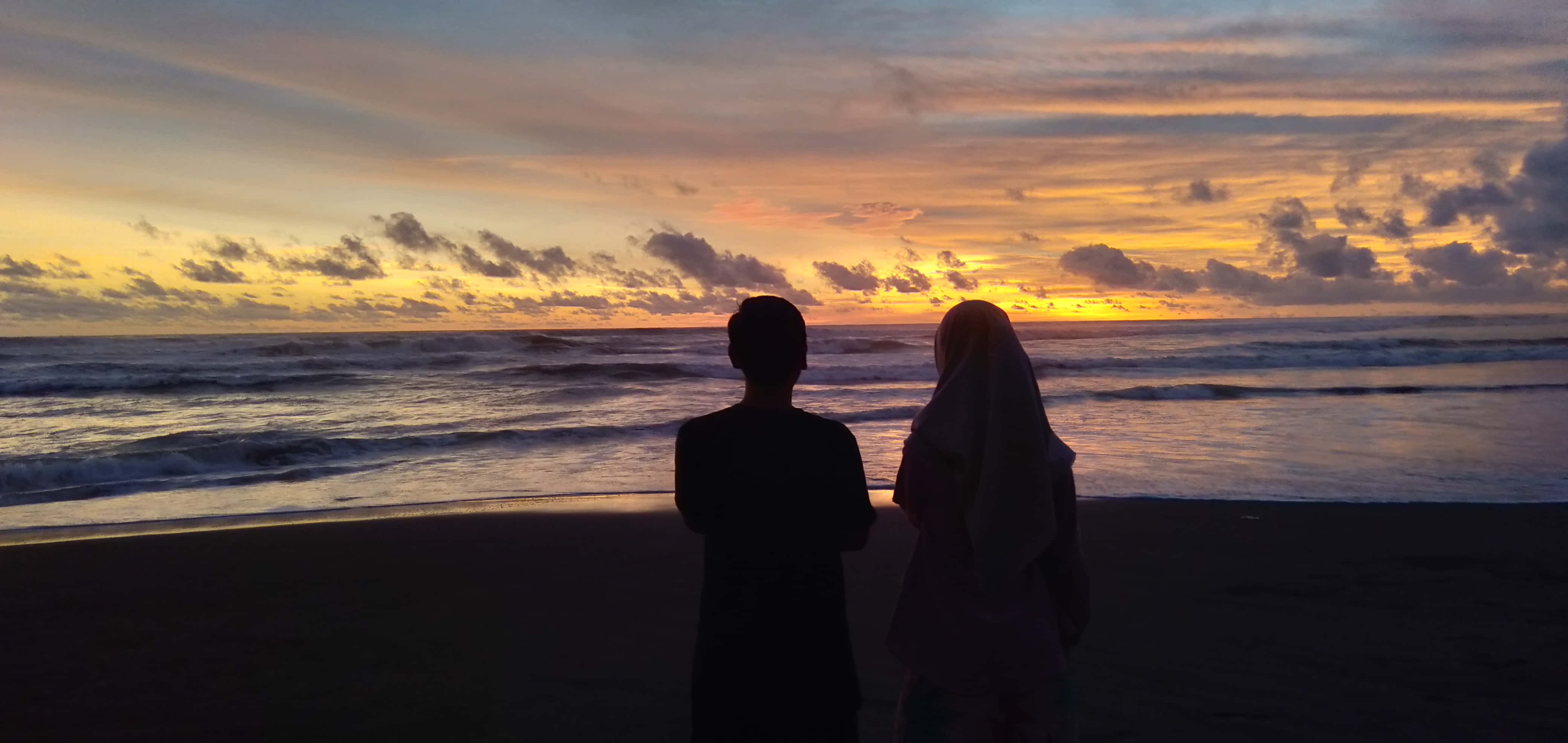 Romance 桌布 壁紙 — 一對情侶的剪影站在沙灘上，觀賞夕陽將天空染成橙色、黃色和紫色，映照在海浪之上。