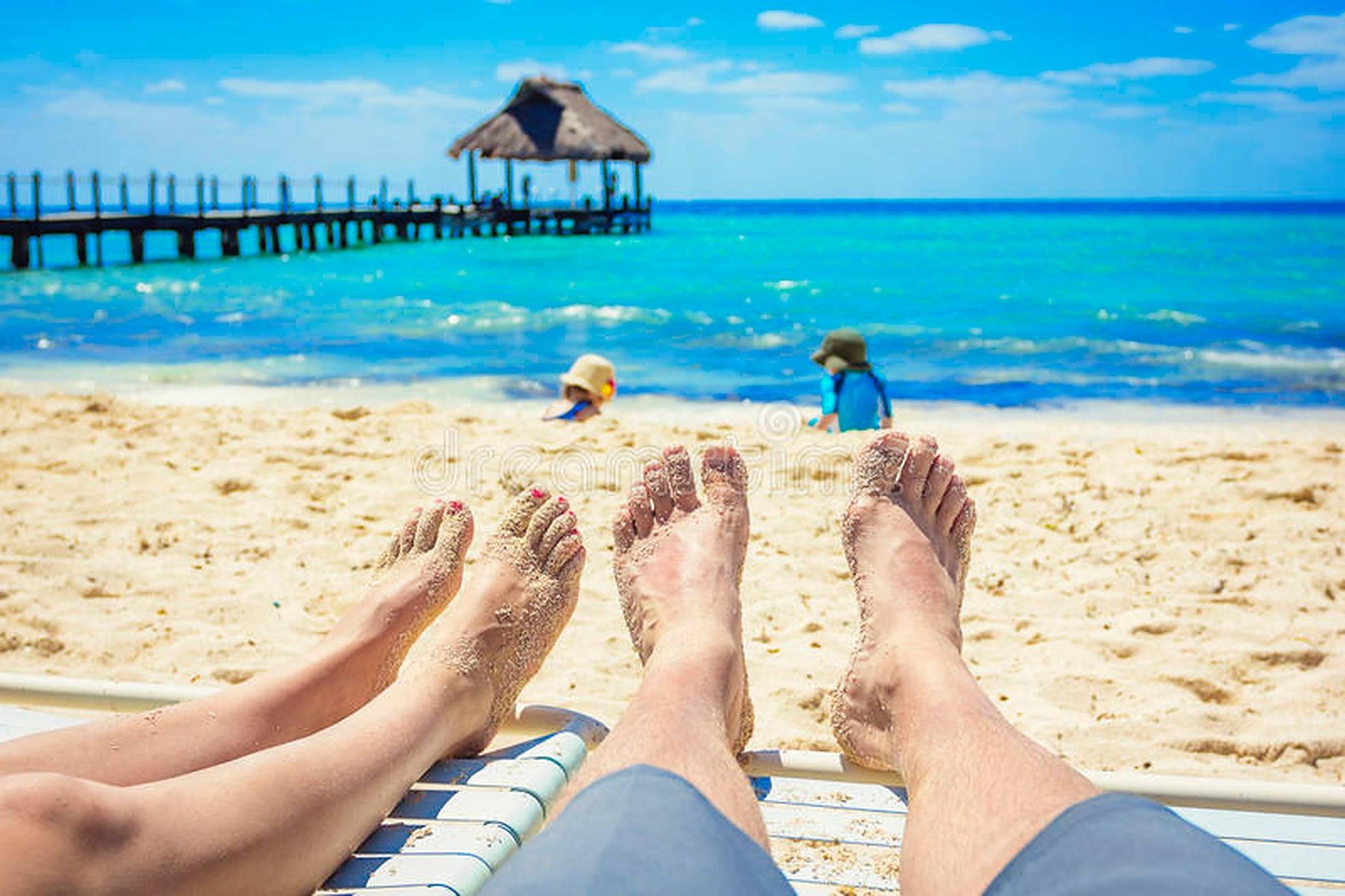 Beach Vacation 桌布 壁紙 — 一對情侶的沙腳在海灘椅上放鬆，孩子們在明亮的藍天下在碼頭附近玩耍。